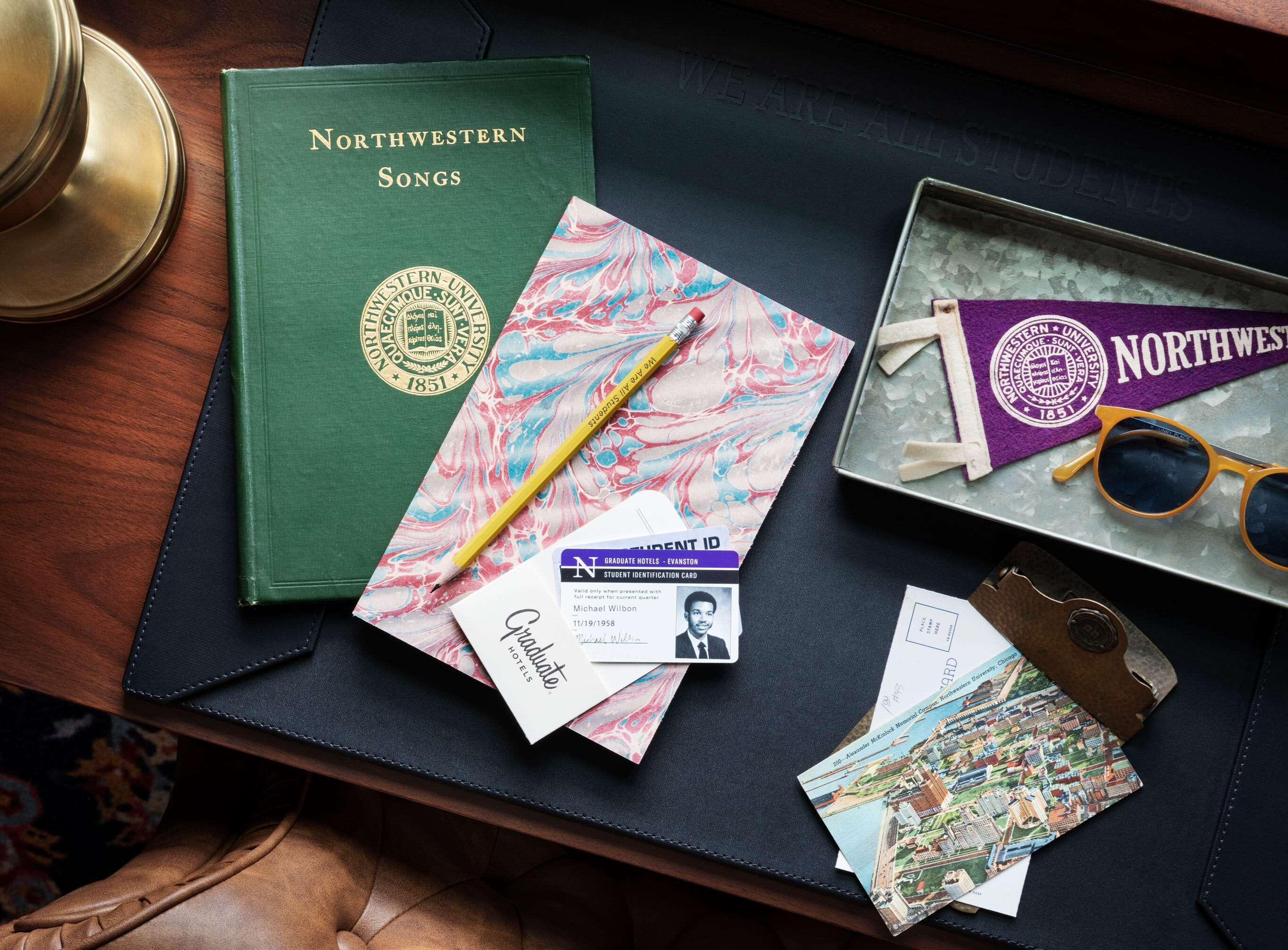 A desk with a green book titled 'Northwestern Songs', a marbled paper, a yellow pencil, a graduate ID card, a pair of sunglasses, and a vintage keychain.