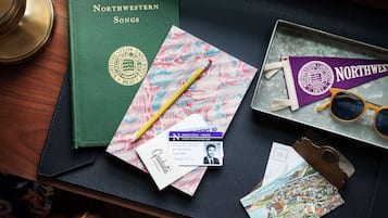 A desk with a green book titled 'Northwestern Songs', a marbled paper, a yellow pencil, a graduate ID card, a pair of sunglasses, and a vintage keychain.