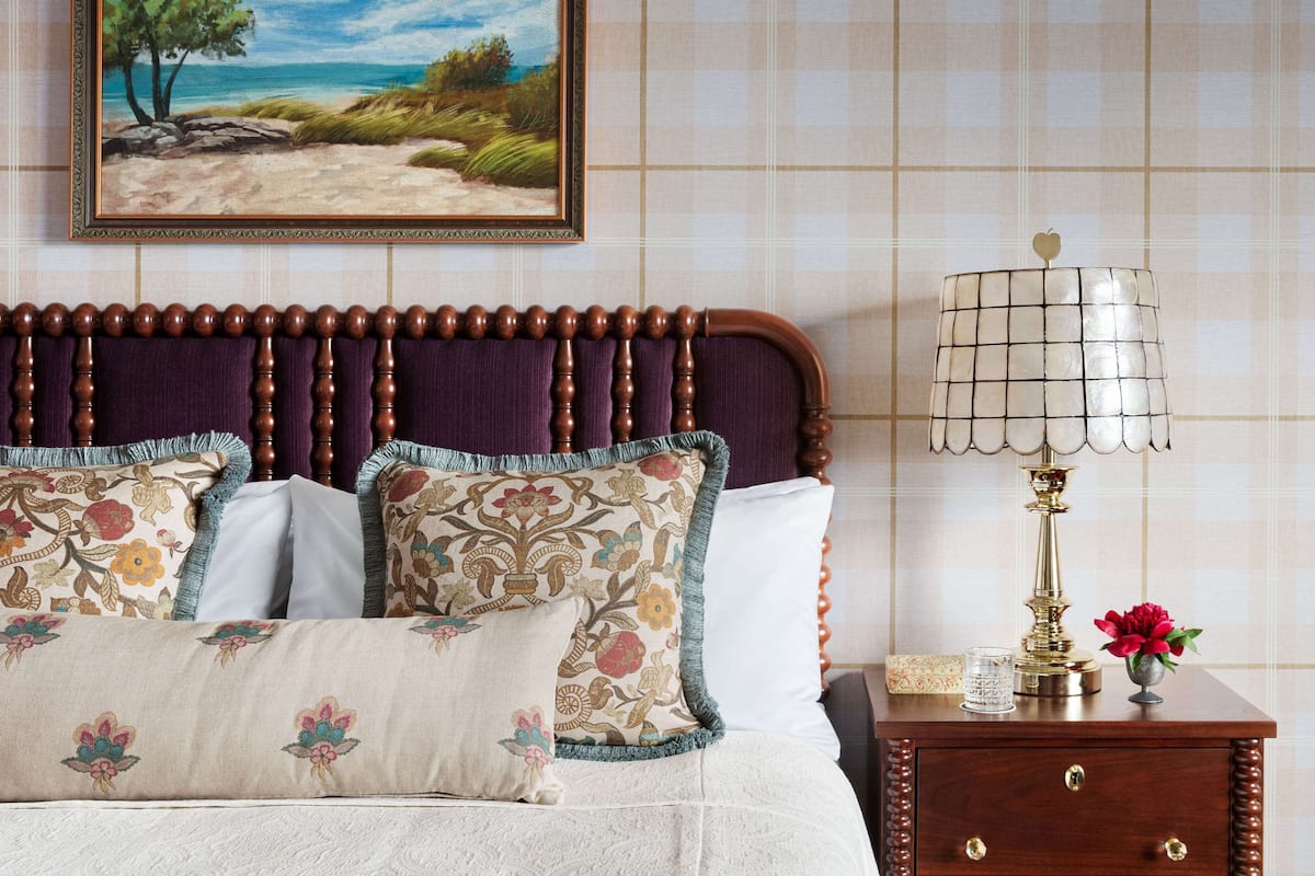 Un dormitorio con cabecera de madera, almohadas estampadas con motivos florales, un cuadro que muestra una playa, una lámpara de noche y una mesita de noche con un florero.