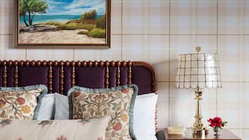 A bedroom with a wooden headboard, floral pillows, a painting of a beach scene, a bedside lamp, and a nightstand with a vase of flowers.