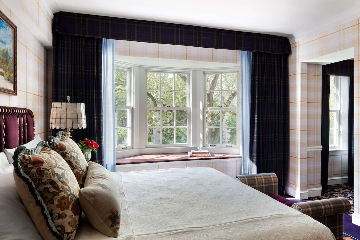 Un dormitorio con una cama grande, un asiento junto a la ventana, una lámpara y vistas a árboles.