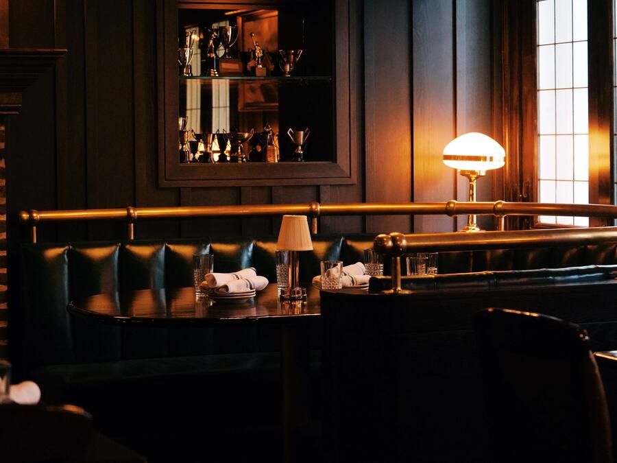 A dimly lit restaurant with leather booths, a table with a lamp, and a mirror reflecting trophies.