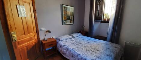 Quarto com porta de madeira, mesa de cabeceira com abajur, cama com roupa de cama estampada, quadro na parede e janela com cortinas.