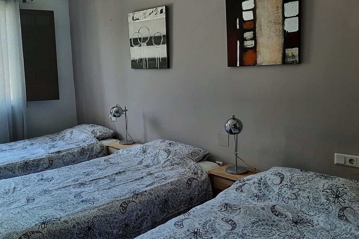 A room with three beds, each with patterned bedding, two bedside tables with lamps, and a window with curtains.