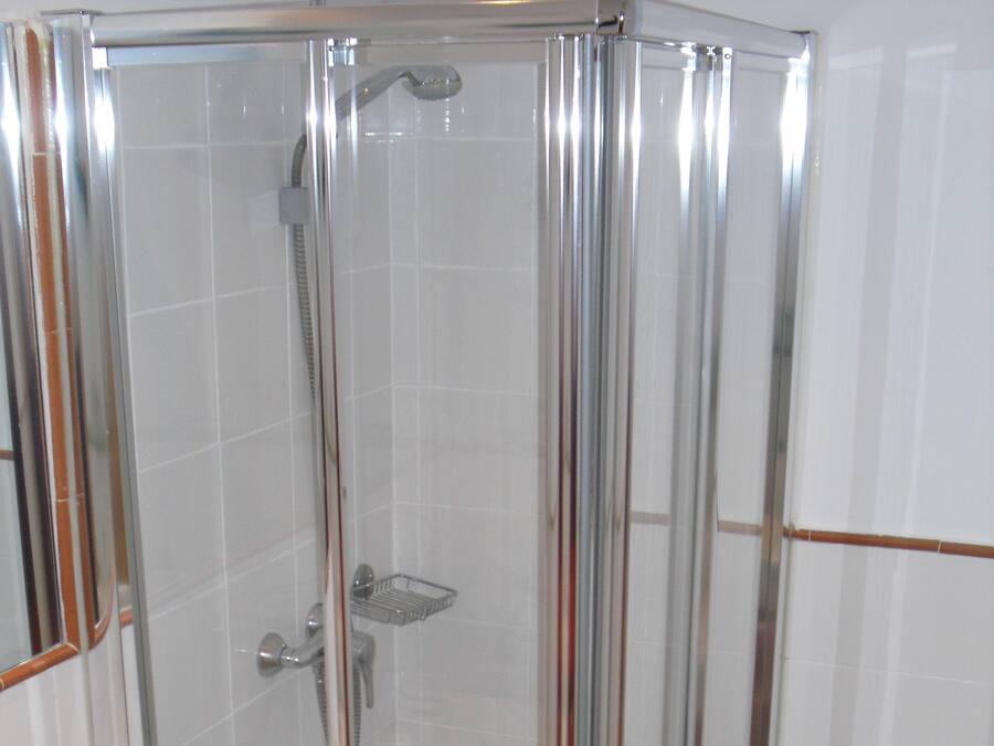 Ducha con cerramiento de vidrio y estructura de cromo, paredes de azulejos blancos y un inodoro a la derecha.