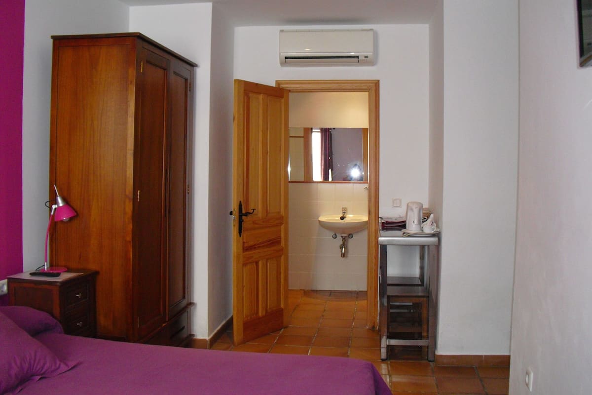 A bedroom with a bed, a wardrobe, a bedside lamp, and a bathroom with a sink and mirror.