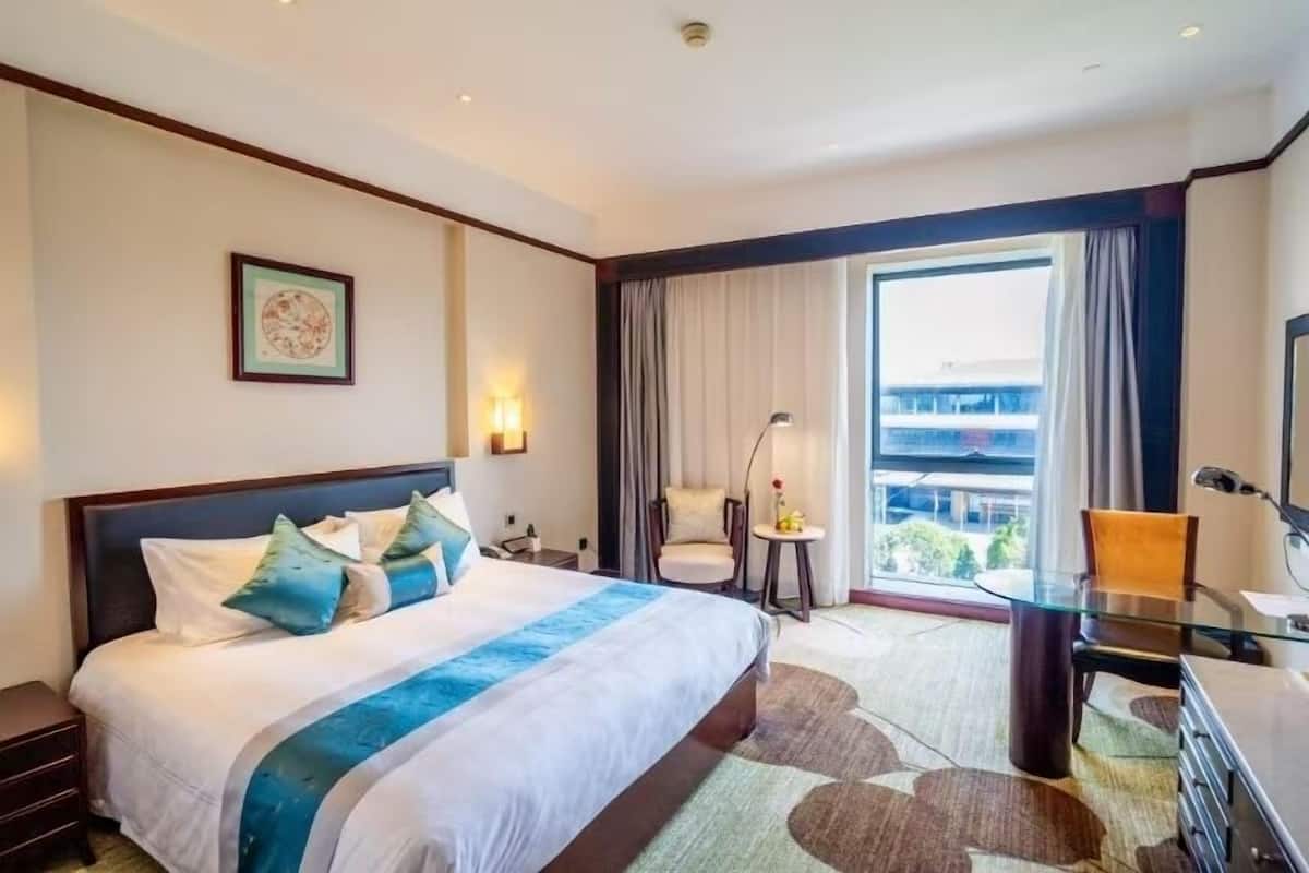 A hotel room with a large bed, a desk, a chair, a window with curtains, and a view of a building.