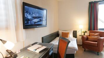Een hotelkamer met een bed, een bureau met lamp, een stoel, een aan de muur bevestigde televisie en een raam met gordijnen.