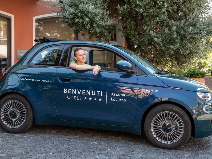Un coche azul oscuro y compacto con la marca 'BENVENUTI HOTELS', estacionado en una calle adoquinada.