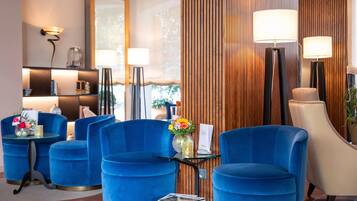 A modern hotel lobby with blue armchairs, a glass table with a vase of flowers, and a wooden panel wall.