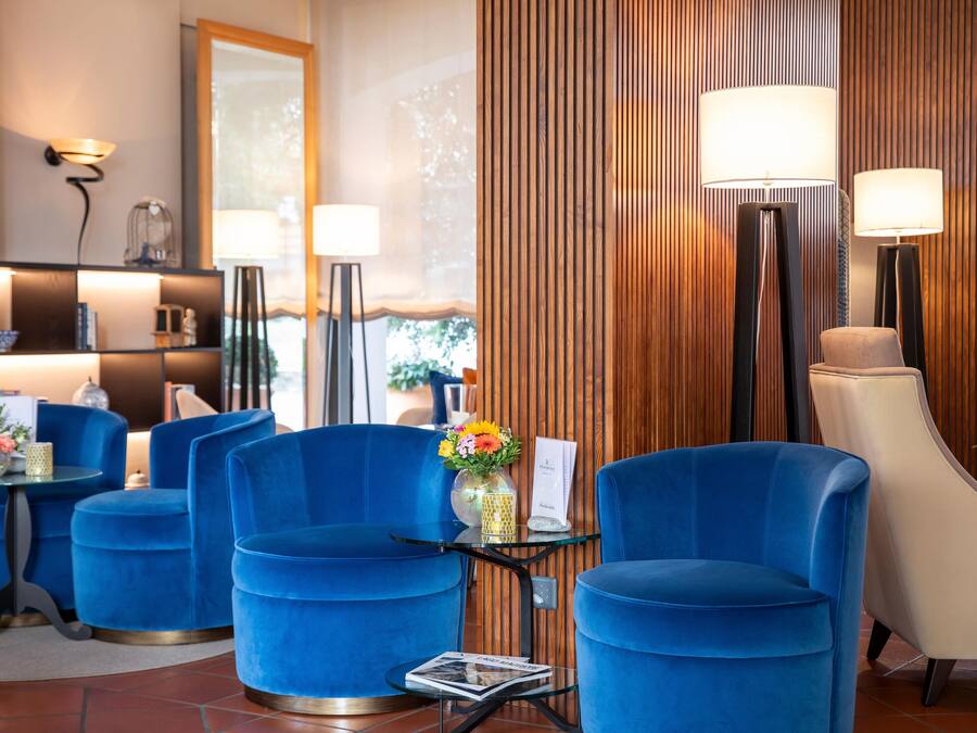 Un hall d’hôtel moderne avec des fauteuils bleus, une table en verre ornée d’un vase de fleurs et un mur revêtu de panneaux en bois.