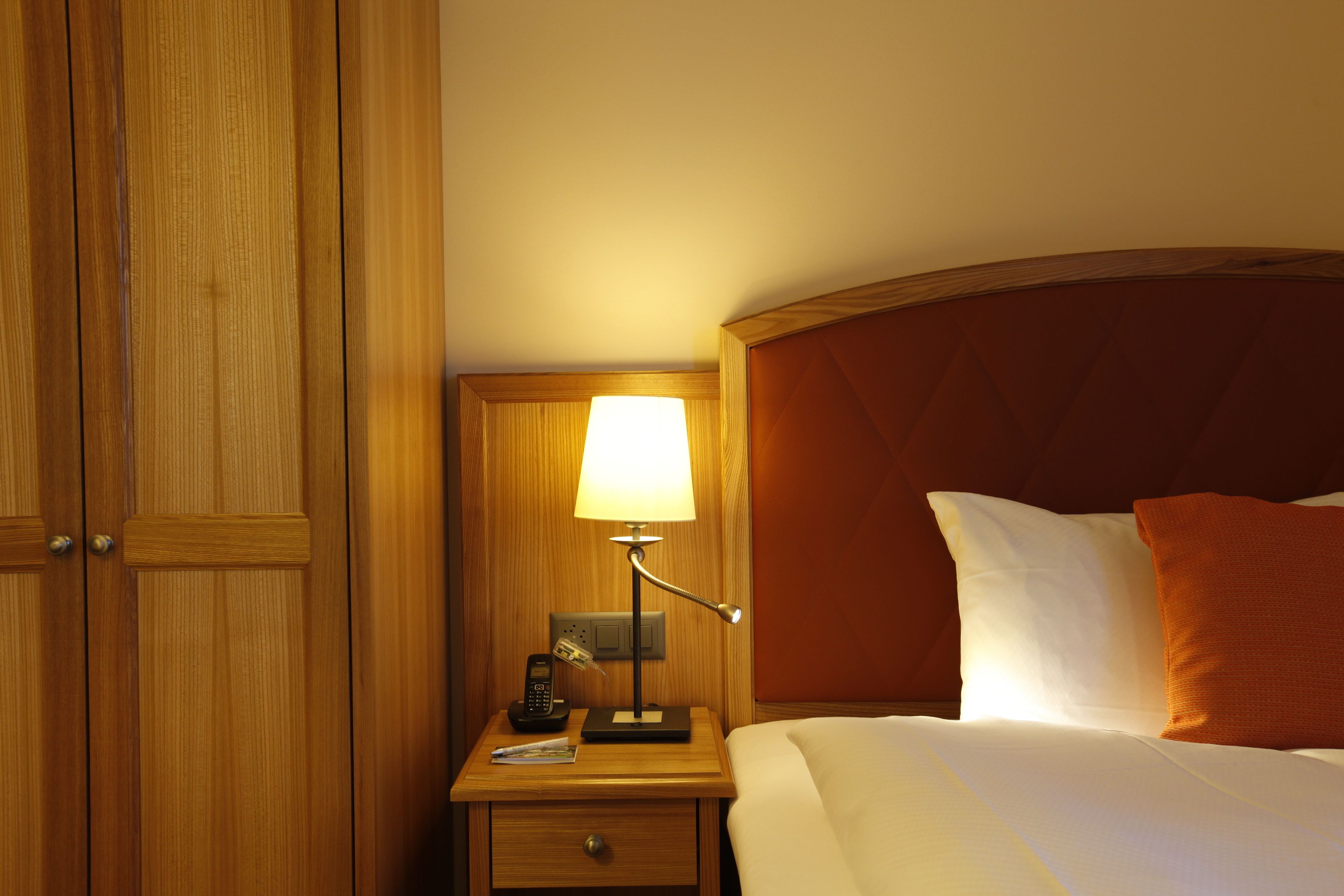 Une chambre d’hôtel avec une armoire en bois, une table de chevet avec une lampe, un téléphone et un lit bien fait, agrémenté d’oreillers.