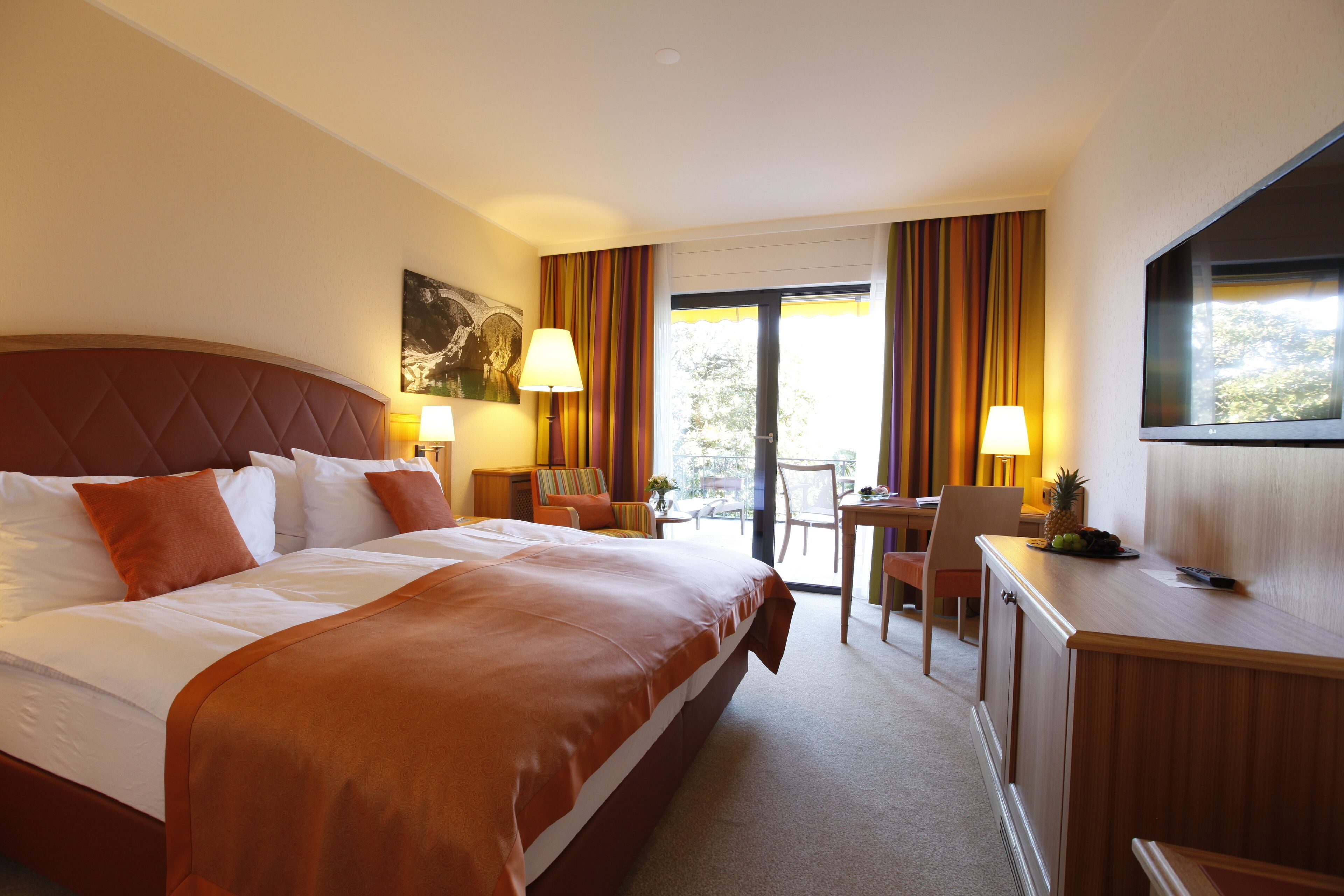 A hotel room with a large bed, a desk, a chair, a TV, and a balcony with curtains.