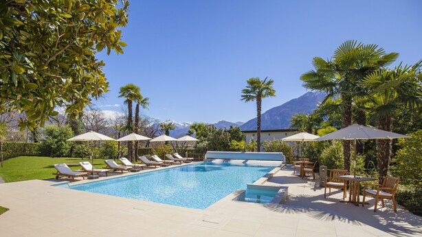 A pool area with lounge chairs and umbrellas, surrounded by greenery and mountains in the background. - Boutique-Hotel REMORINO (Minusio)