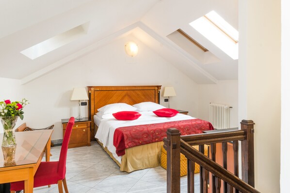 A bedroom with a wooden bed, two bedside lamps, a red throw blanket, a red chair, a wooden desk with a vase of flowers, and a radiator. - Hotel Leon D´Oro (Prague)