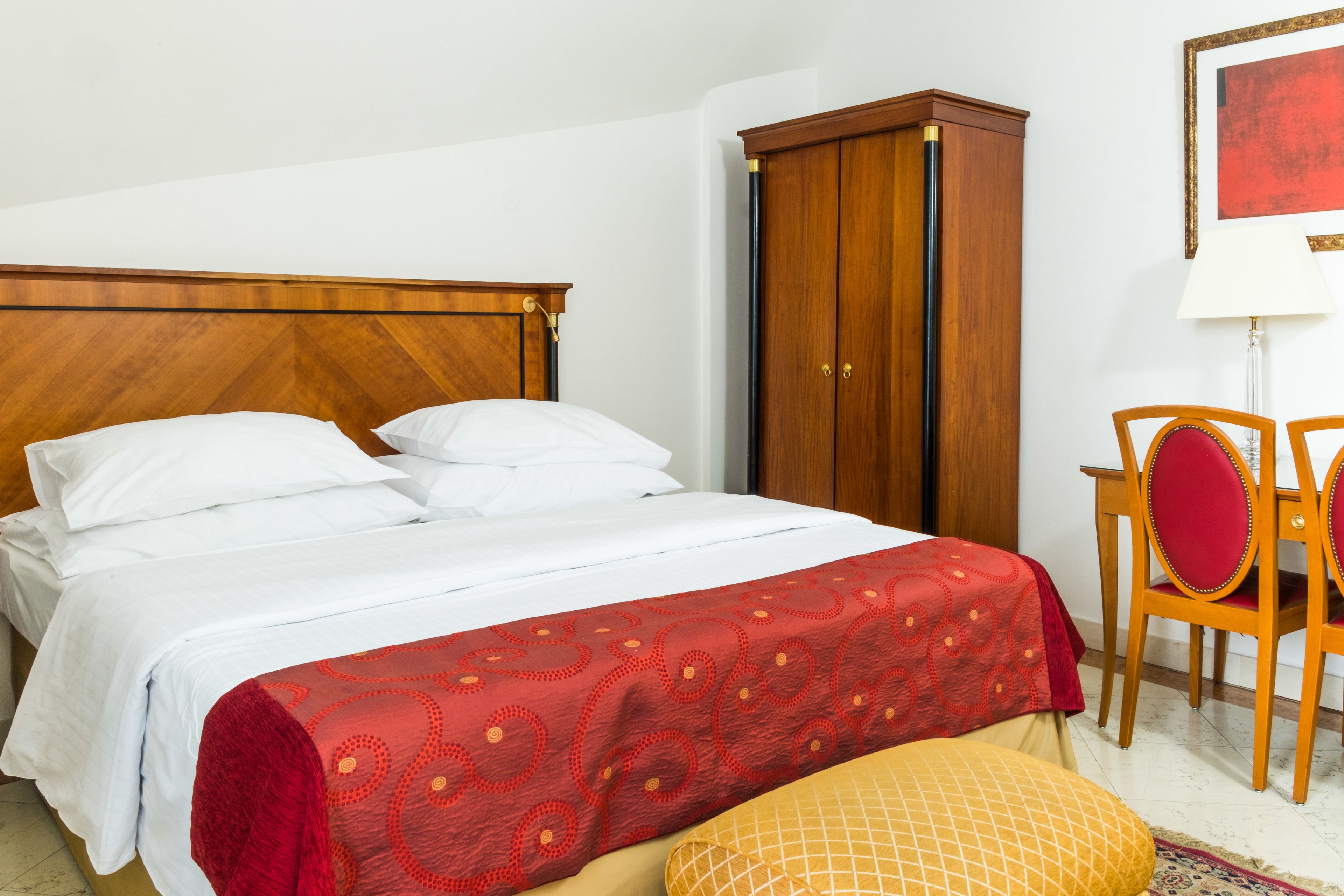 Cama bem arrumada com lençóis brancos, manta vermelha estampada e cabeceira de madeira.