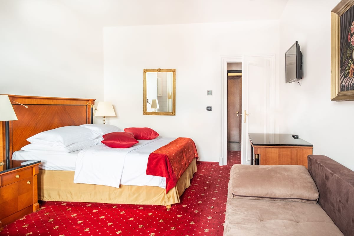 A hotel room with a bed, a bedside table with a lamp, a red carpet, a brown sofa, and a painting on the wall.