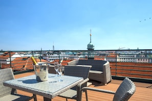 Una terraza en la azotea con muebles de mimbre, una mesa con un cubo de champán y copas, y vistas de la ciudad que incluyen una torre.