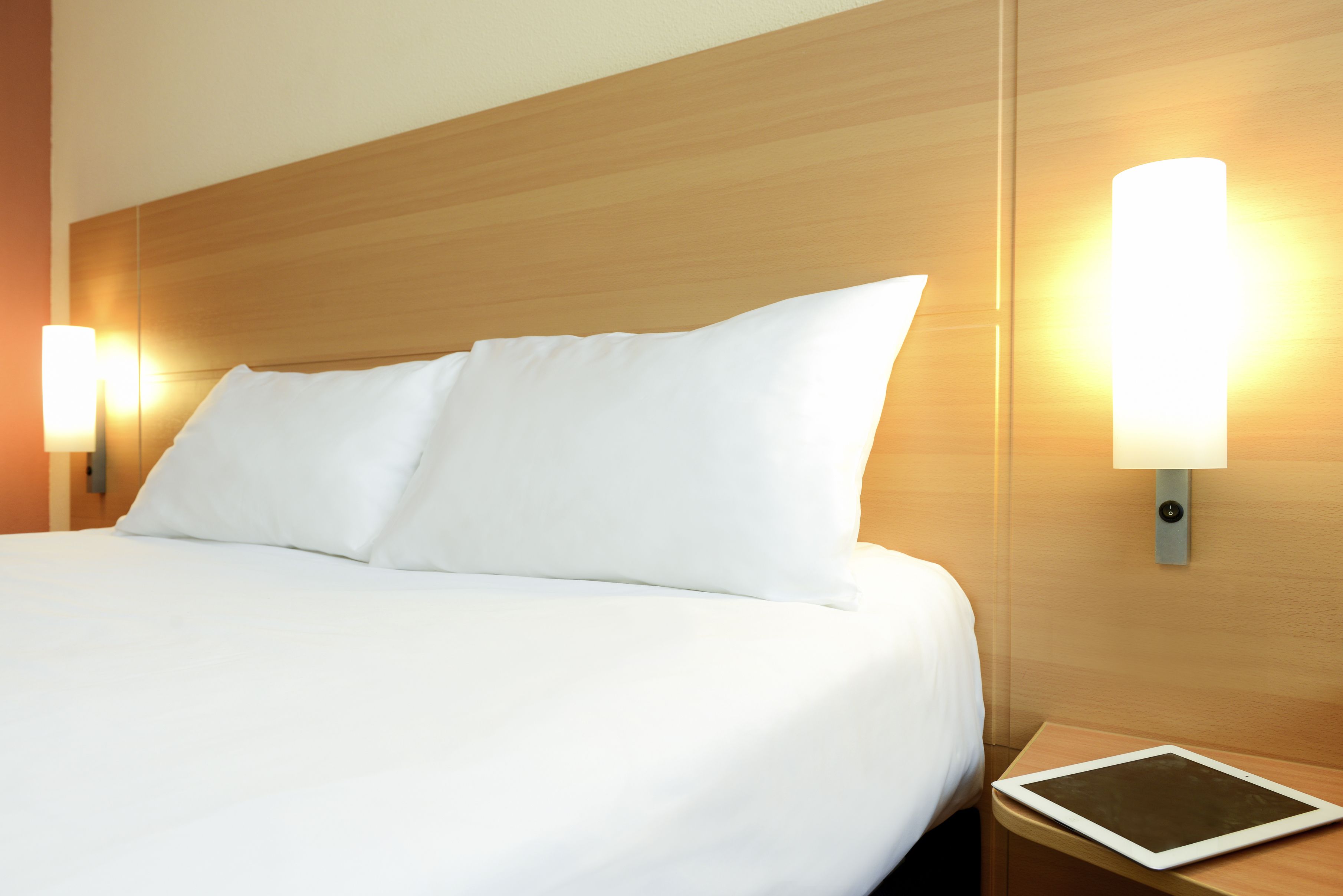 A neatly made bed with white linens, two wall-mounted bedside lamps, and a tablet on the nightstand.
