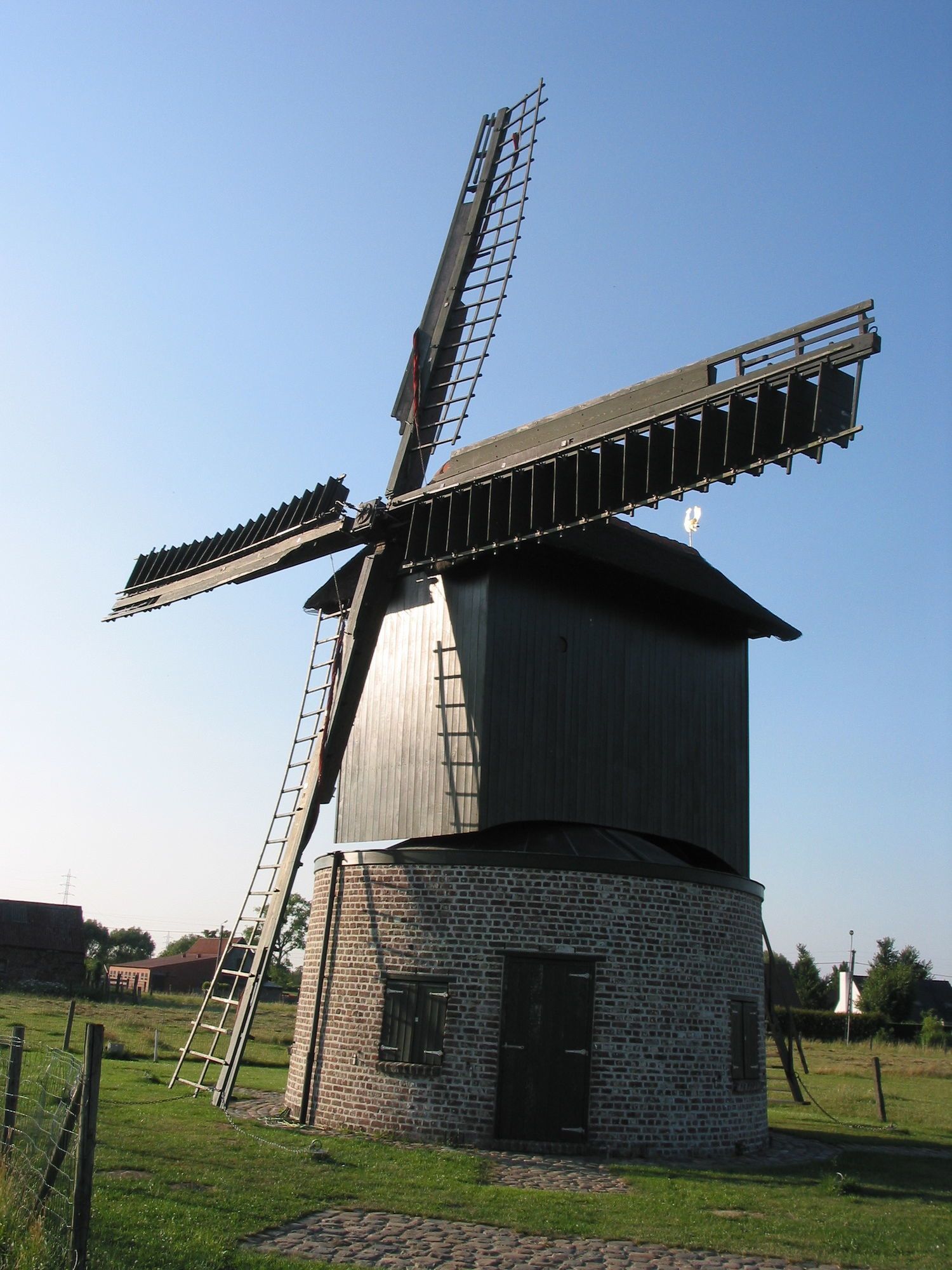Een windmolen met een bakstenen fundering en een metalen constructie, omringd door een gazon en een keienpad.