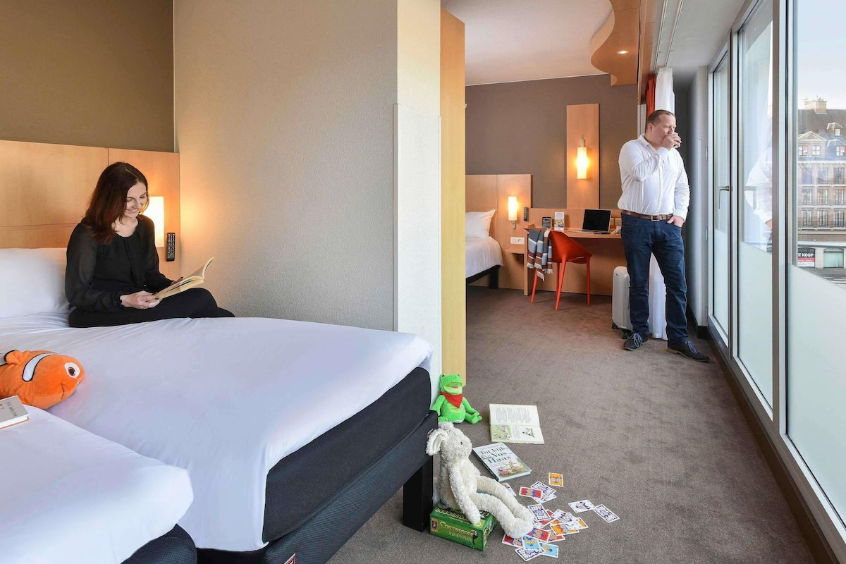 Ein Hotelzimmer mit Bett, Schreibtisch und Blick auf die Stadt durch große Fenster.