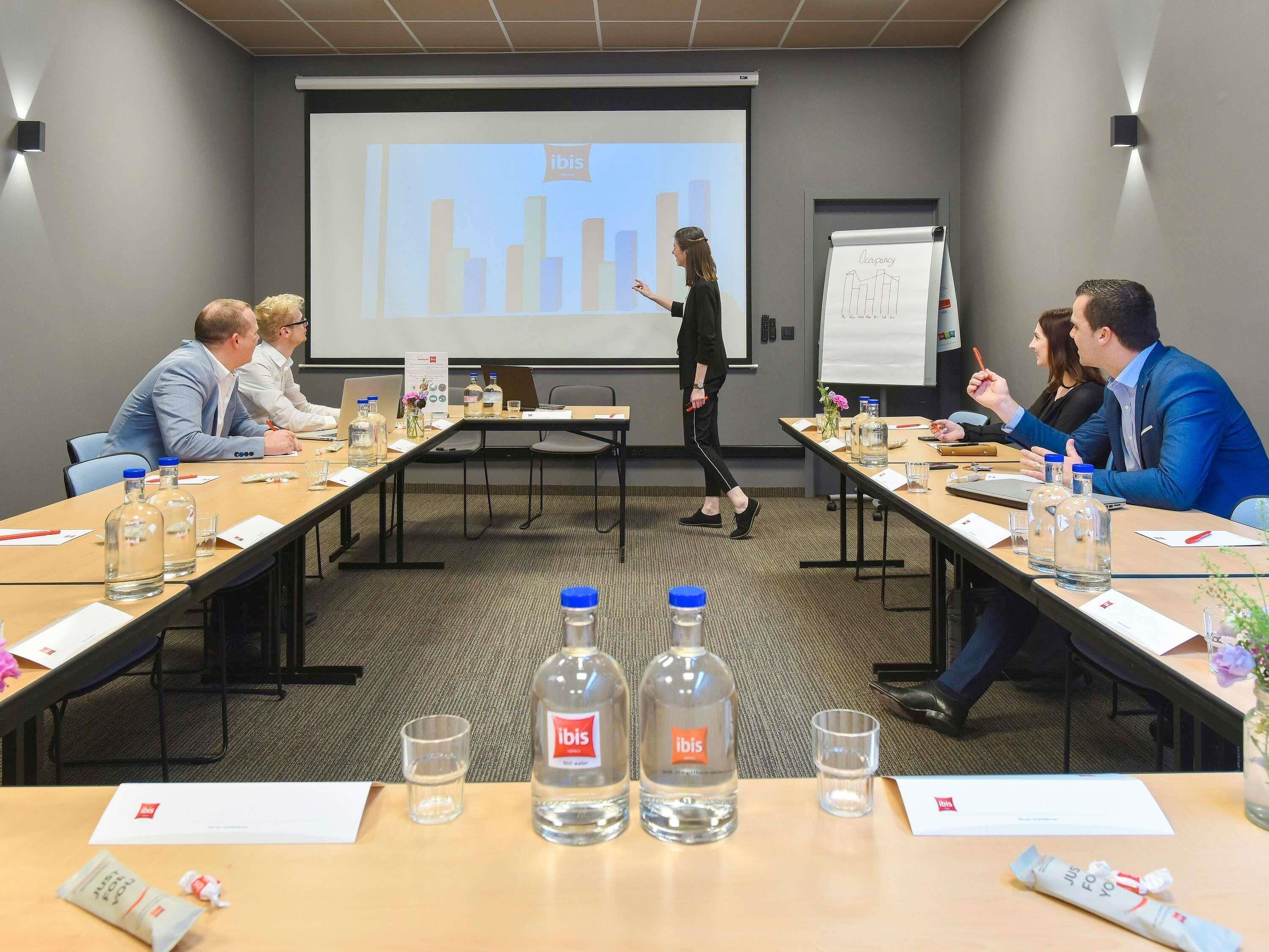 Ein Konferenzraum mit Präsentationsschirm, Whiteboard und einem Tisch mit Wasserflaschen und Dokumenten.