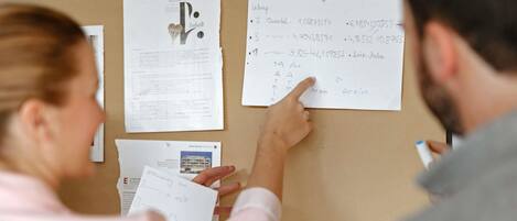 A person pointing at a bulletin board with papers pinned on it, including one with architectural plans and another with handwritten notes.