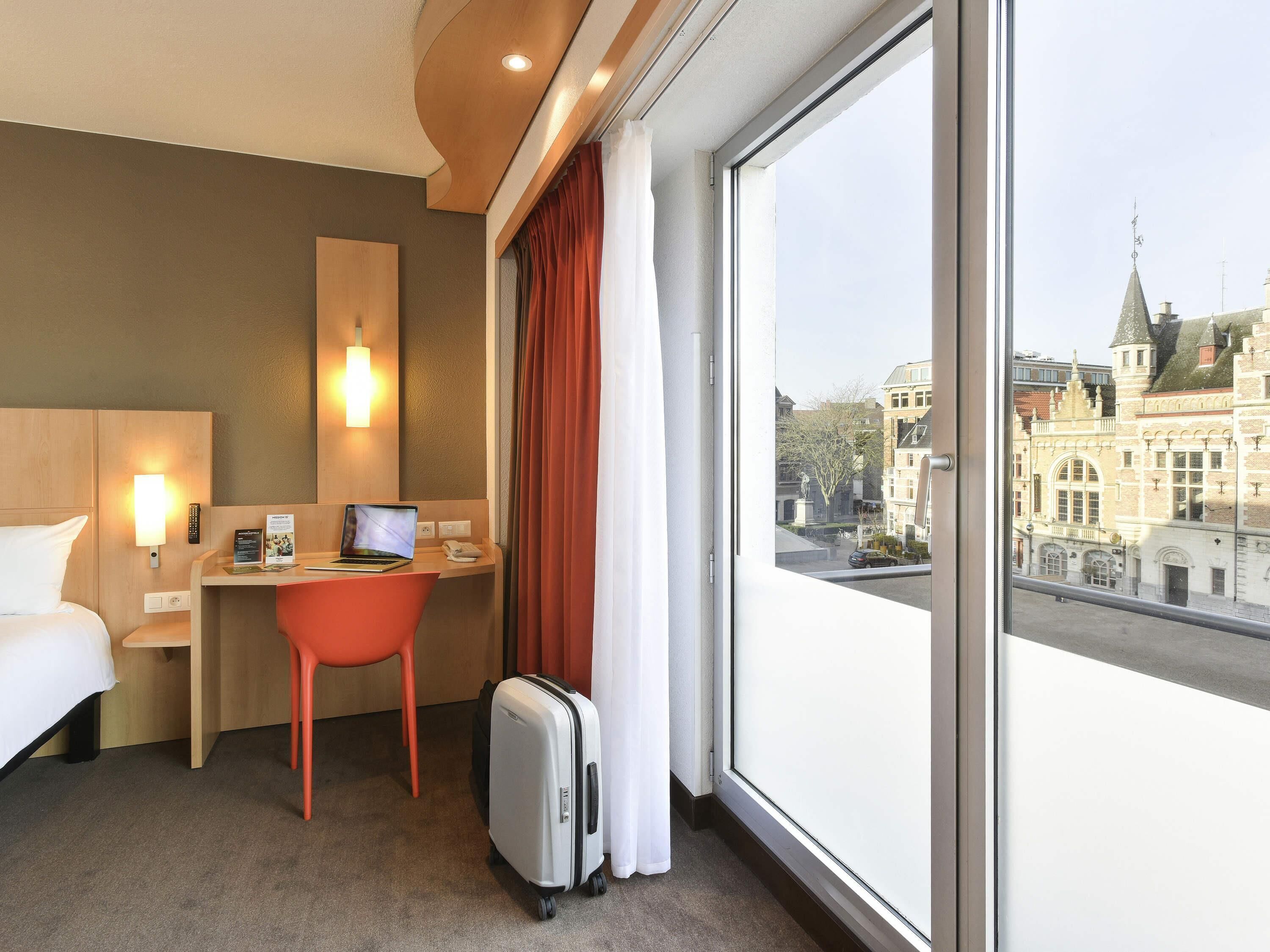 A hotel room with a bed, desk, chair, laptop, and suitcase.