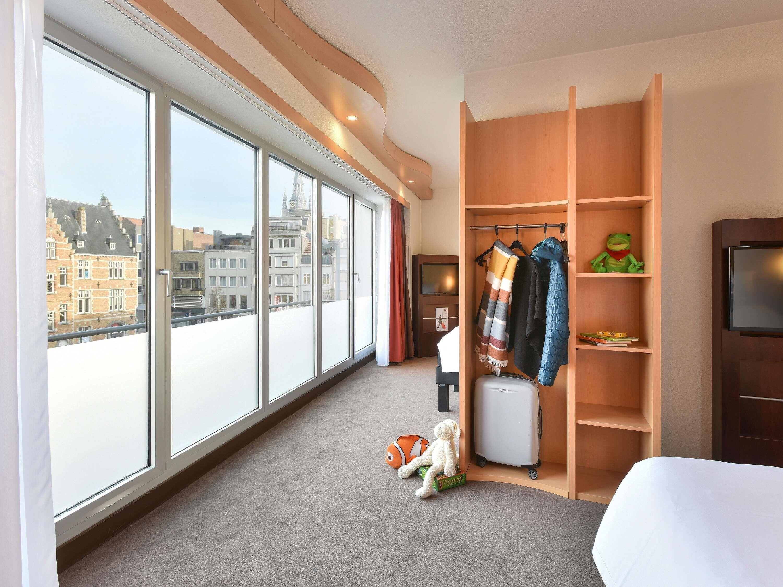 Een hotelkamer met grote ramen, een kast met hangende kleding, een koffer en een klein tafeltje met een speeltje.