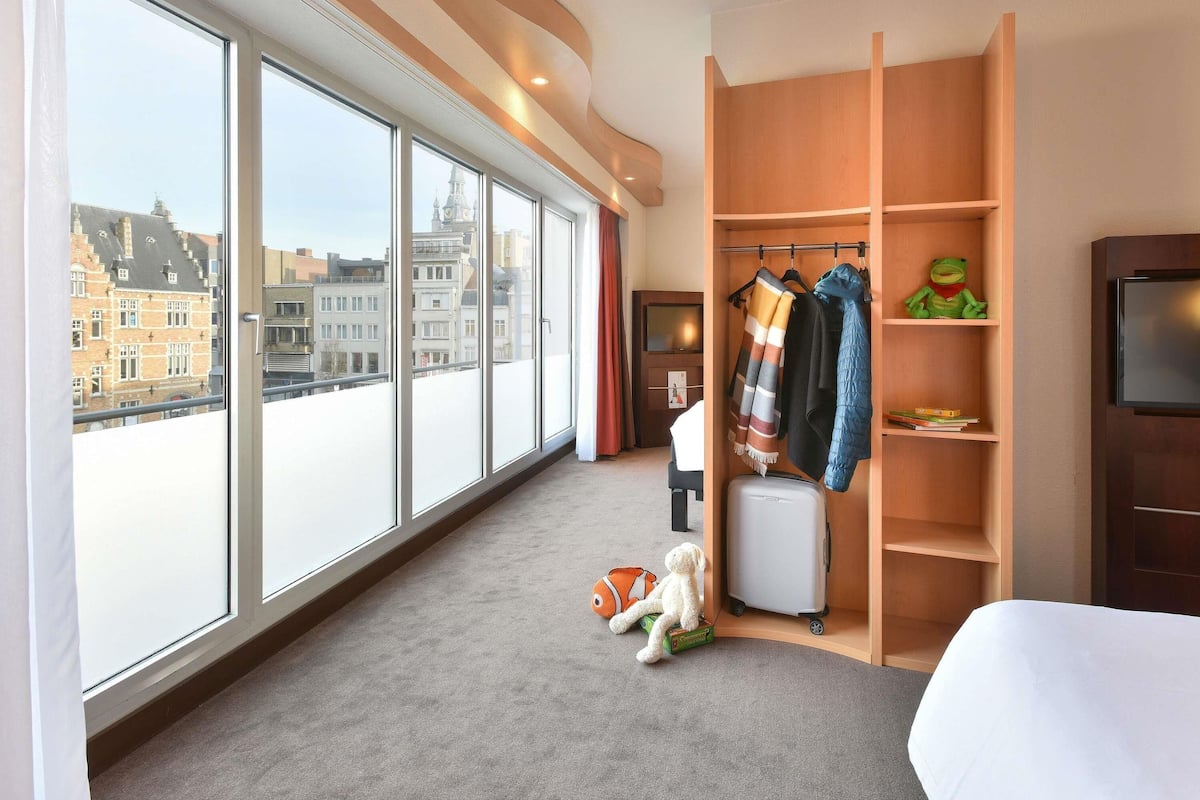 Una habitación de hotel con ventanas amplias, un armario con ropa colgada, una maleta y una pequeña mesa con un juguete.