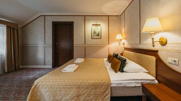 Quarto de hotel com uma cama grande, duas mesas de cabeceira, um criado-mudo, uma porta e uma luminária fixada na parede.