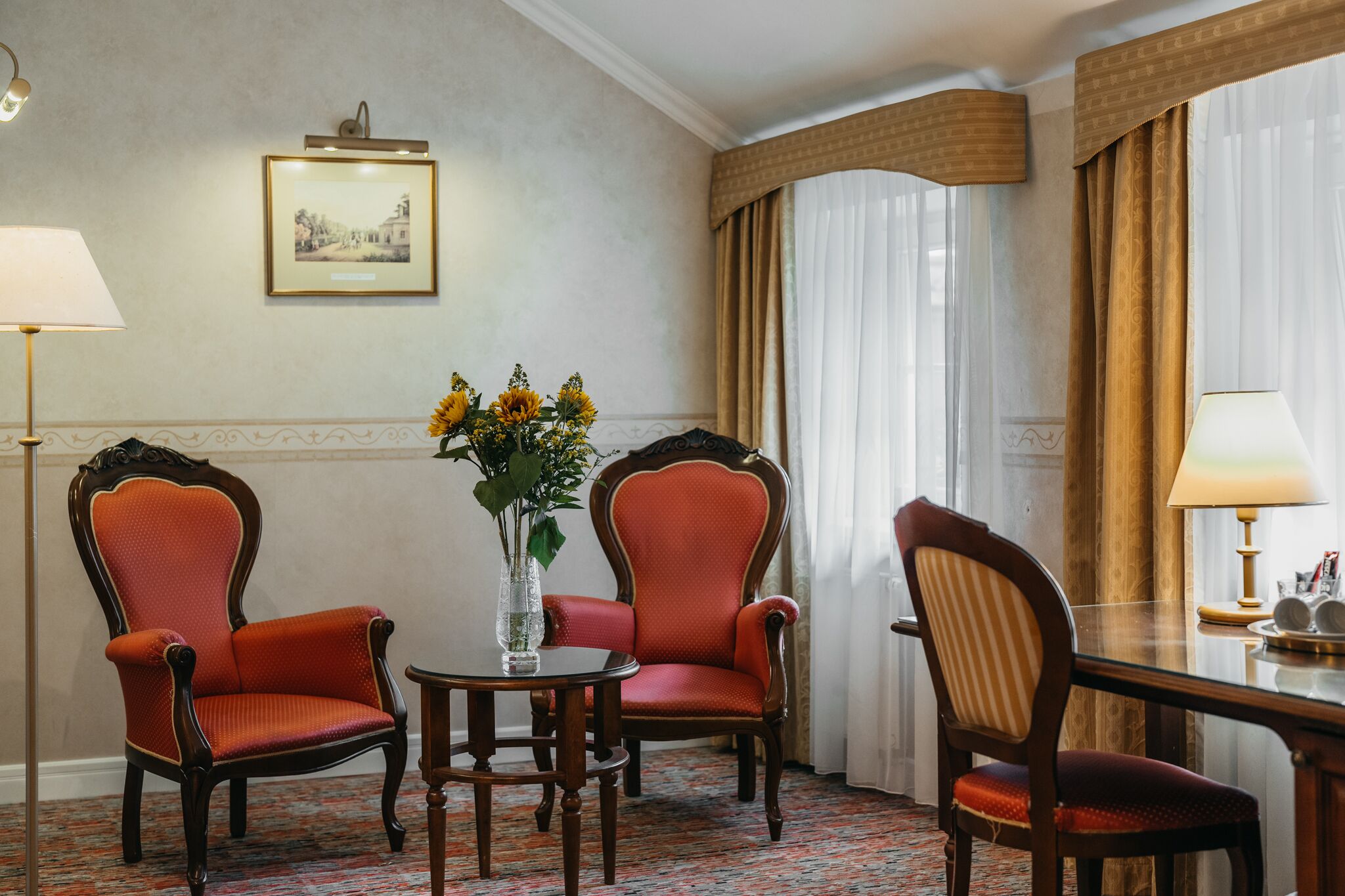 Een hotelkamer met twee fauteuils, een bijzettafeltje, een vaas met bloemen en een lijst met een afbeelding aan de muur.
