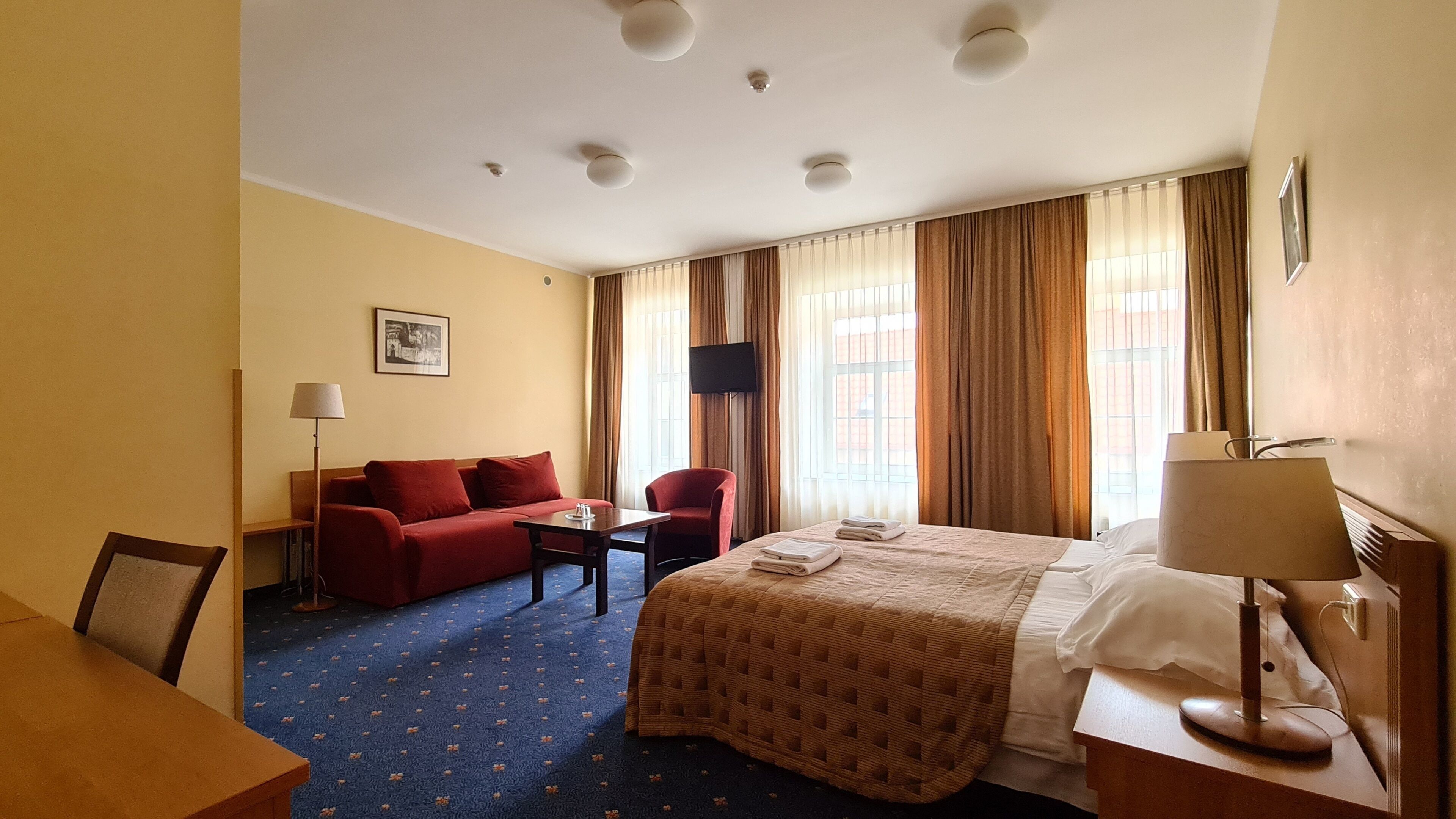 A hotel room with a bed, a red sofa, a desk, a chair, a TV, and a window with curtains.