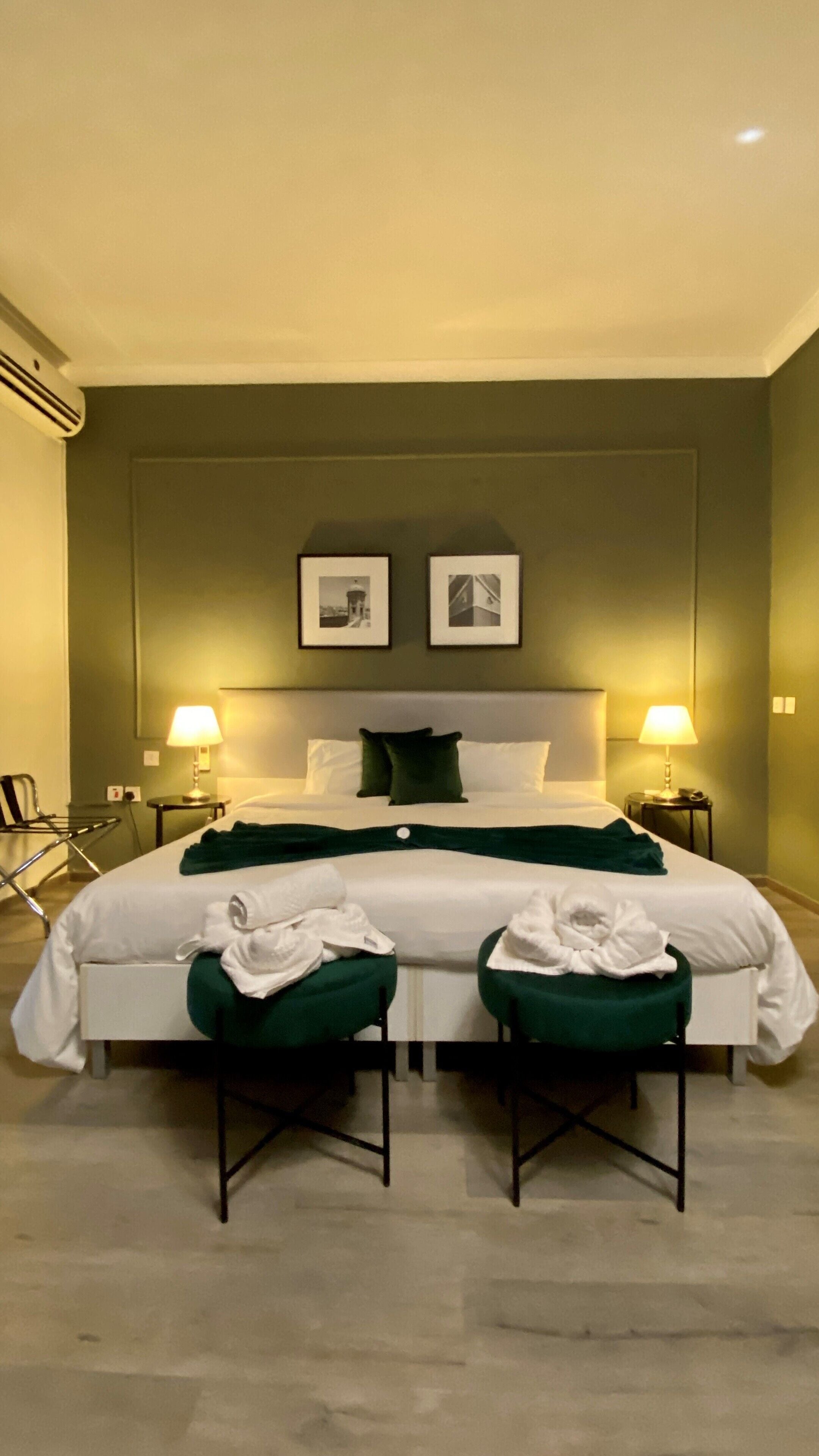 A hotel room with a large bed, two bedside lamps, a headboard with two framed pictures, and two green chairs with towels.