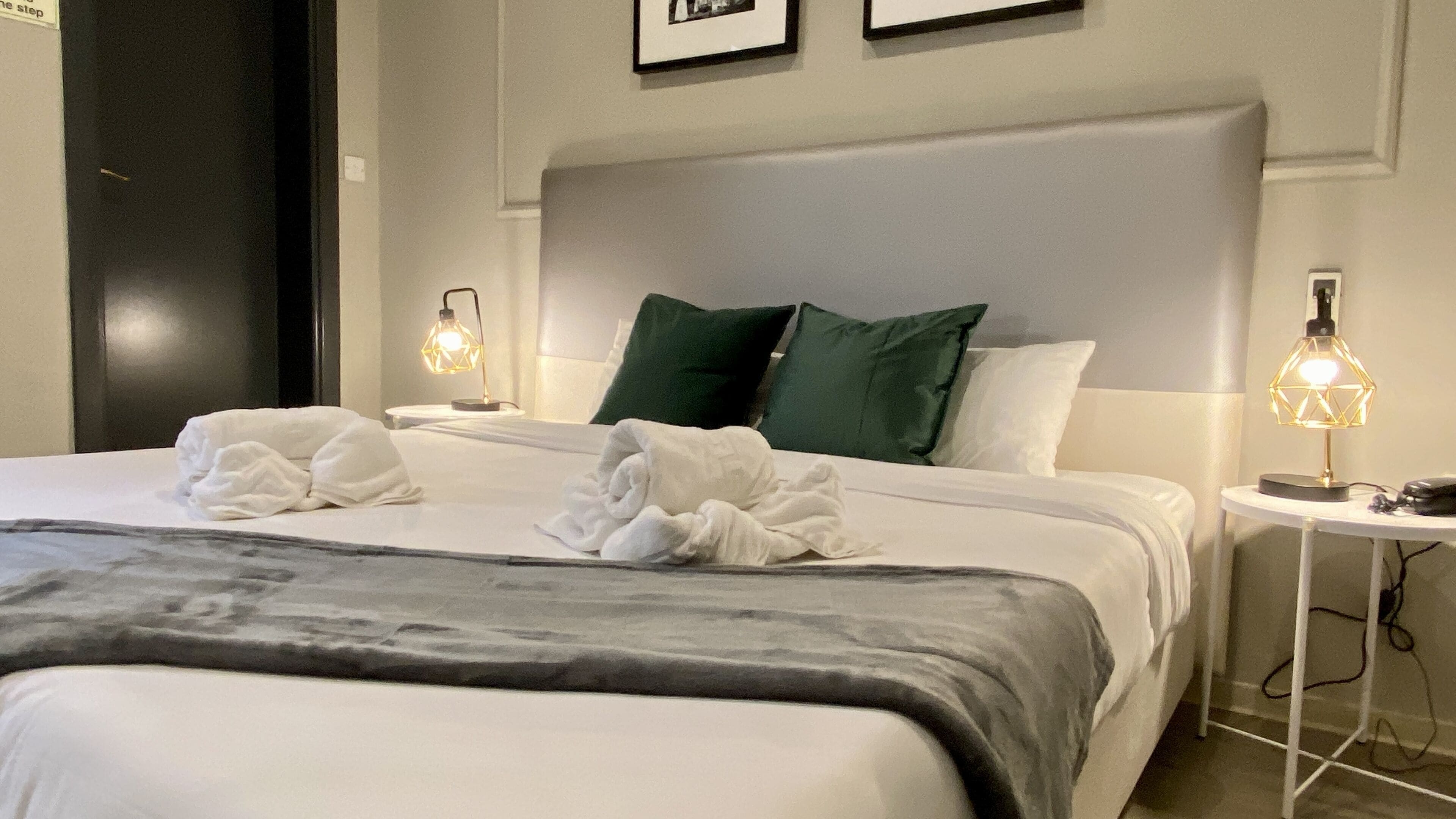 Un lit bien fait, avec du linge de lit blanc et deux serviettes, un plaid gris et deux coussins verts. Deux lampes de chevet aux motifs géométriques sont posées sur les tables de chevet.