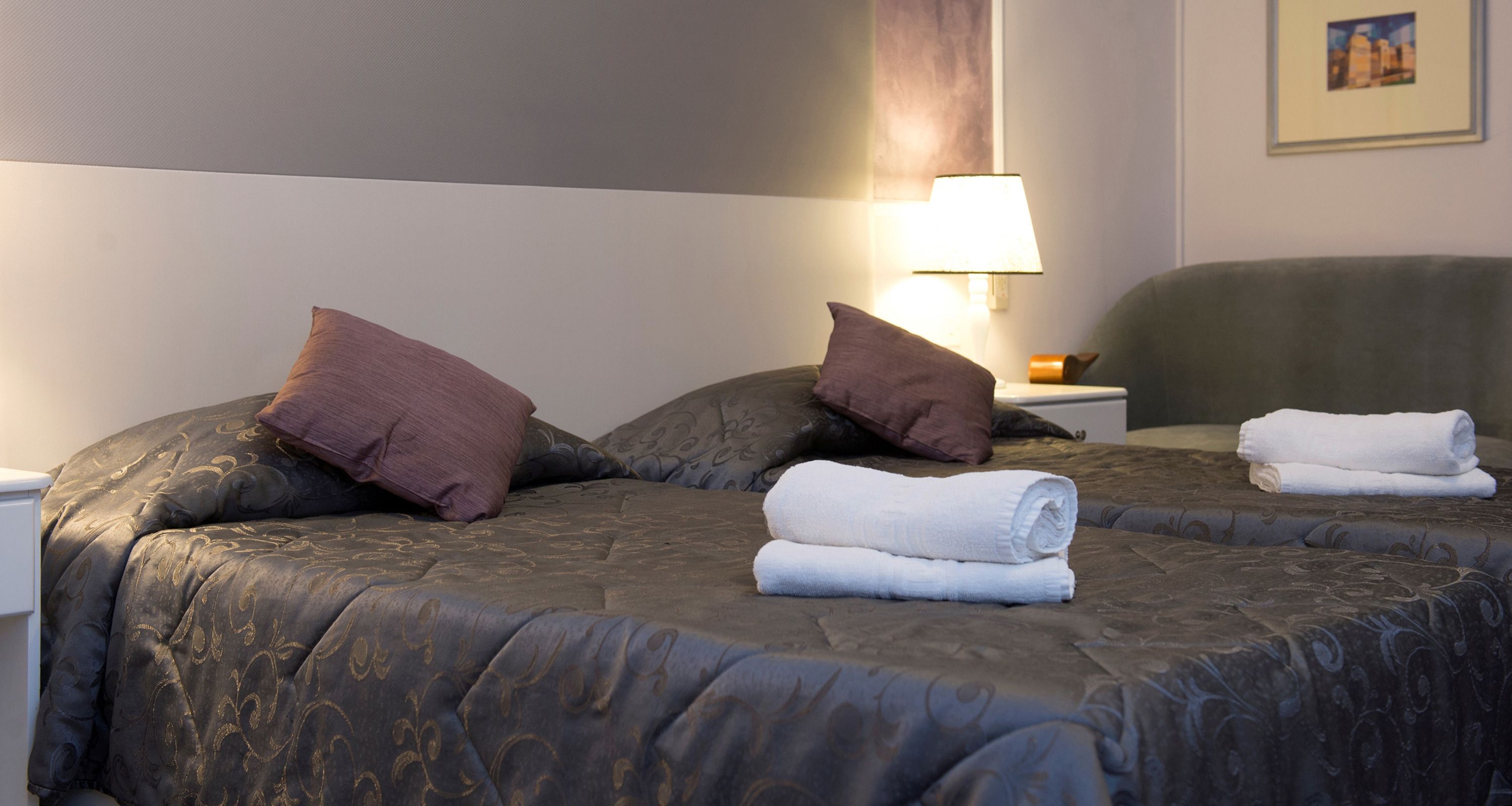 Quarto de hotel com duas camas, cada uma com colcha escura estampada, almofadas e uma toalha branca dobrada. É visível uma mesa de cabeceira com uma luminária e um quadro emoldurado na parede.