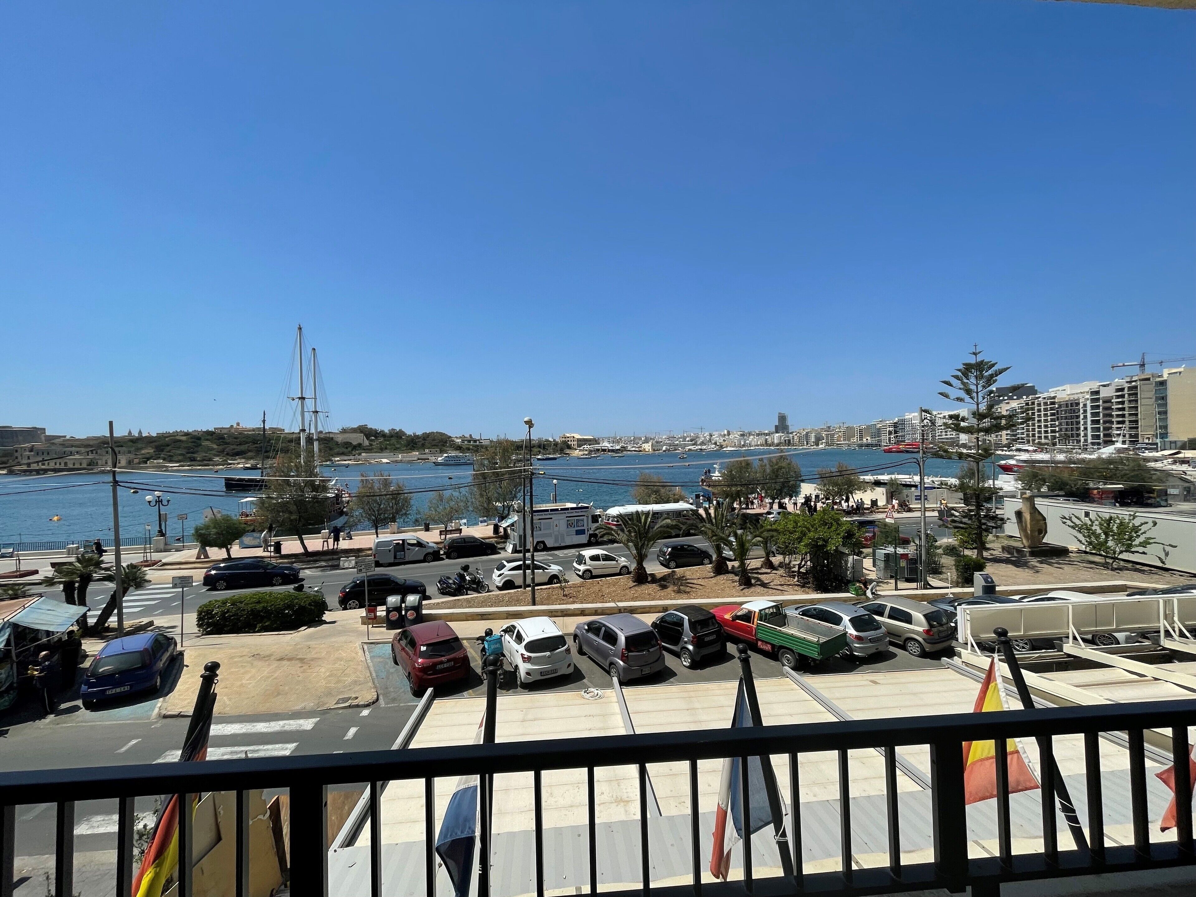 Ein Parkplatz an der Küste mit parkenden Autos, freiem Blick auf das Meer und einer entfernten Stadtlandschaft.