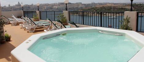 A rooftop pool area with a hot tub, lounge chairs, and a view of the cityscape.