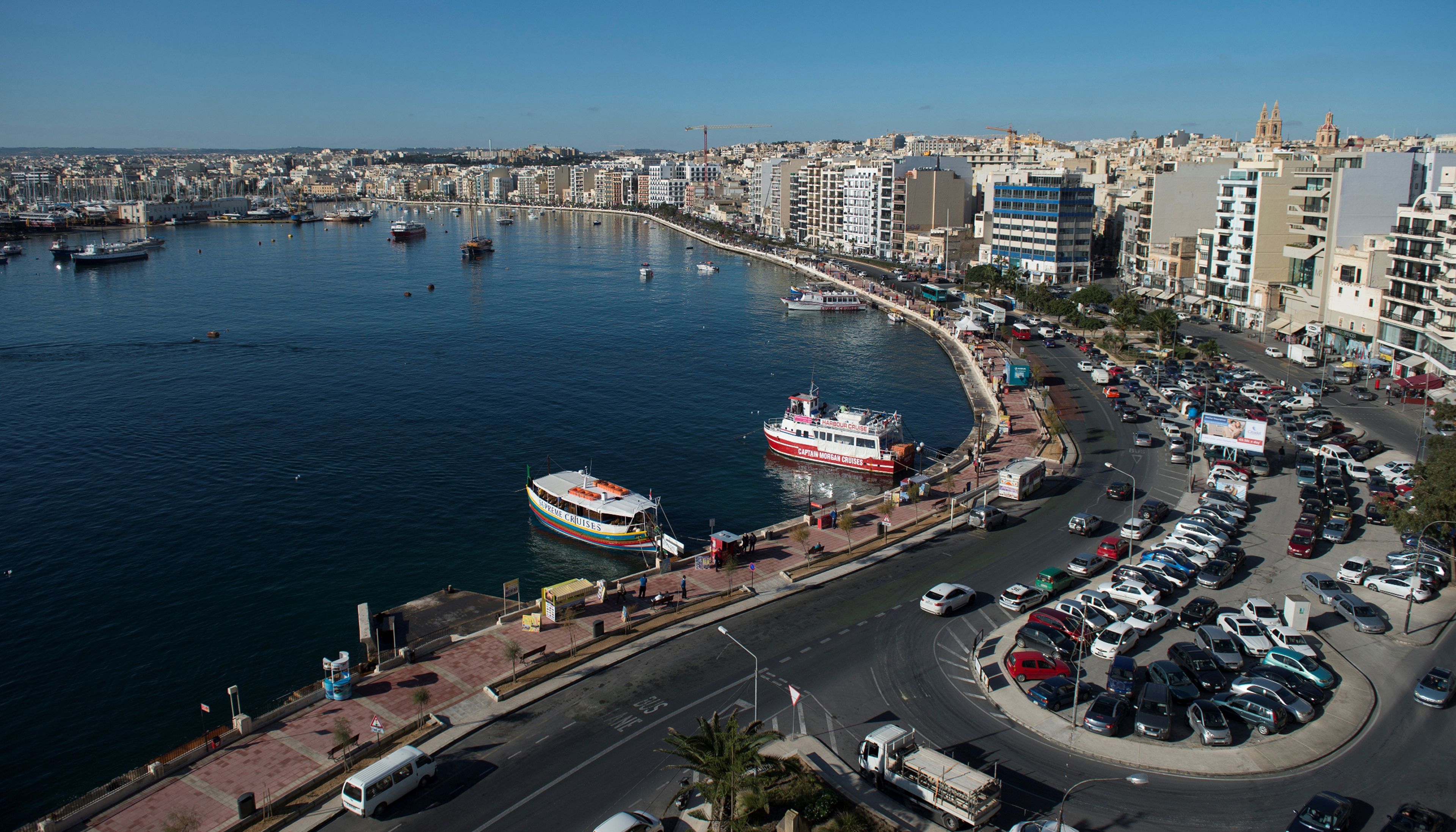 A coastal cityscape with a marina, boats, and a busy road with parked cars.