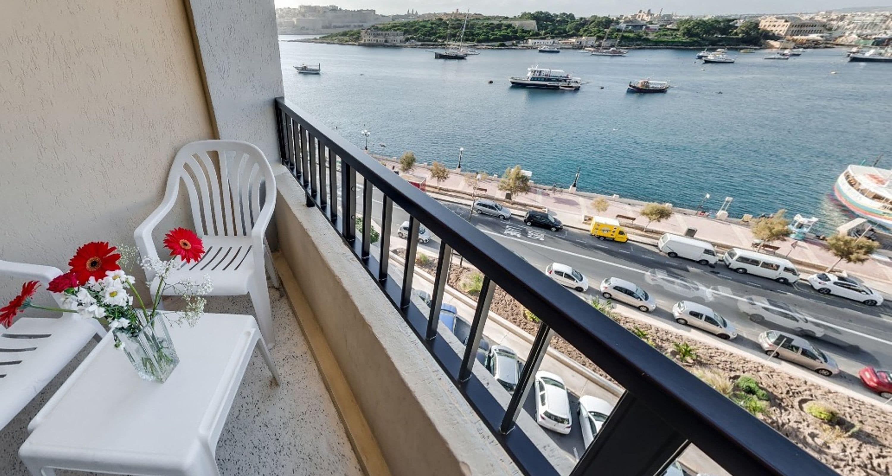 A balcony with a view of a harbor, a white chair, and a table with flowers.