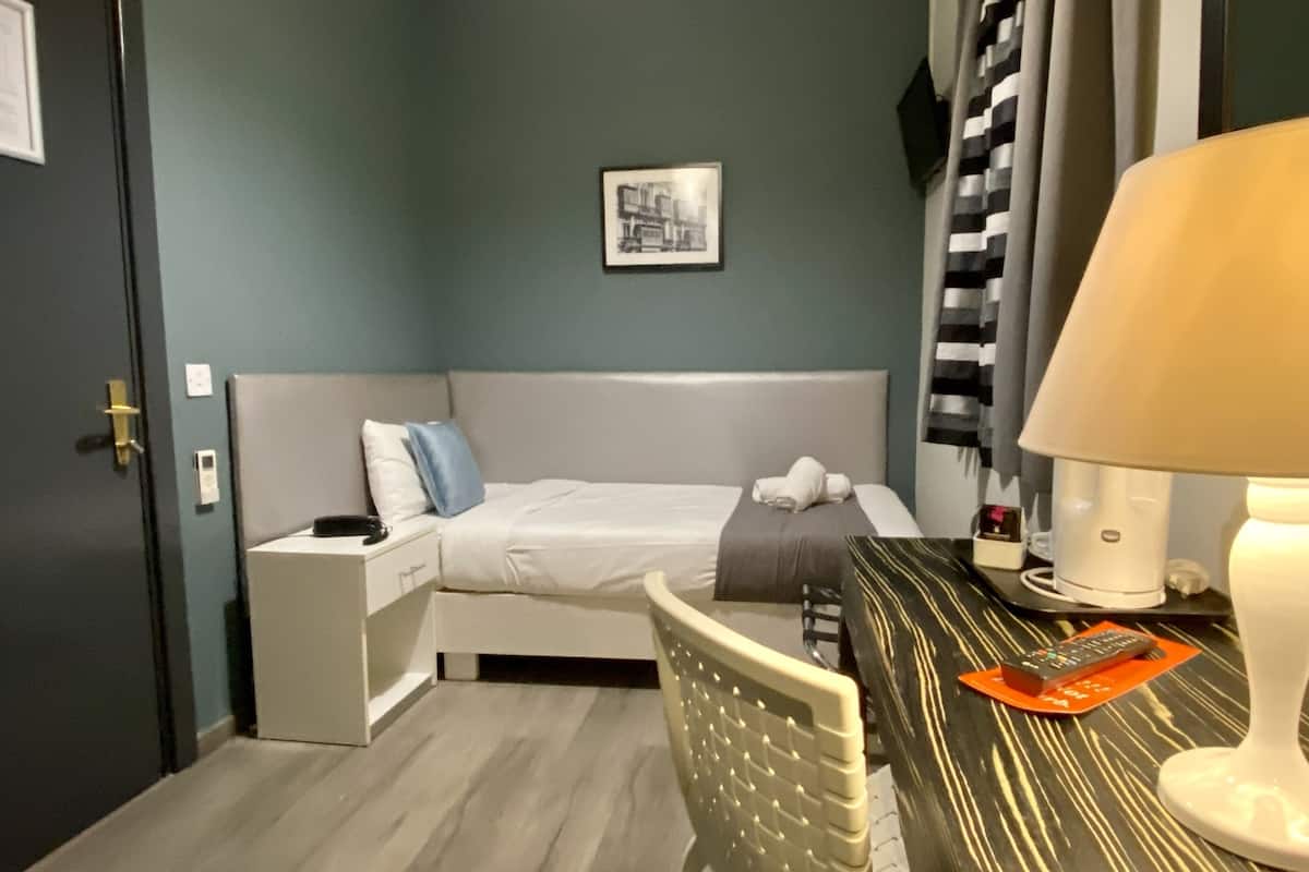 Kompakti hotellihuone, jossa on sänky, yöpöytä, lamppu ja pieni työpöytä tuolilla.