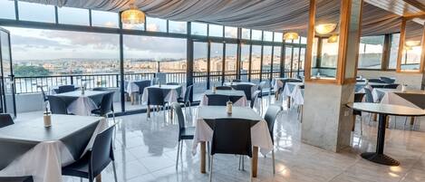 A restaurant with a terrace overlooking a cityscape, featuring tables and chairs, large windows, and hanging light fixtures.