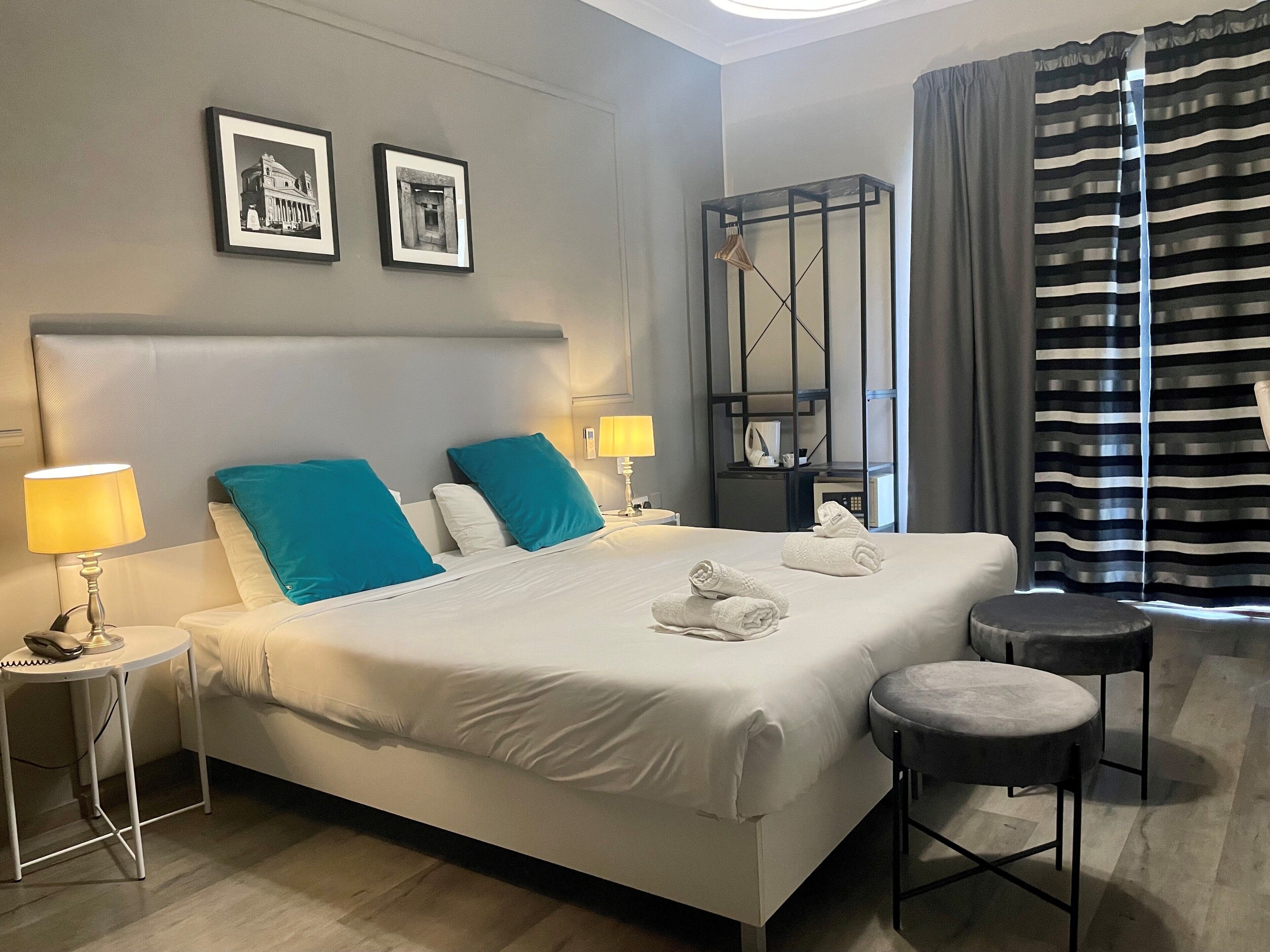 Hotelkamer met een groot bed, twee kussens, een nachtkastje met een lamp, een klein krukje, een spiegel en twee foto’s in lijsten aan de muur.