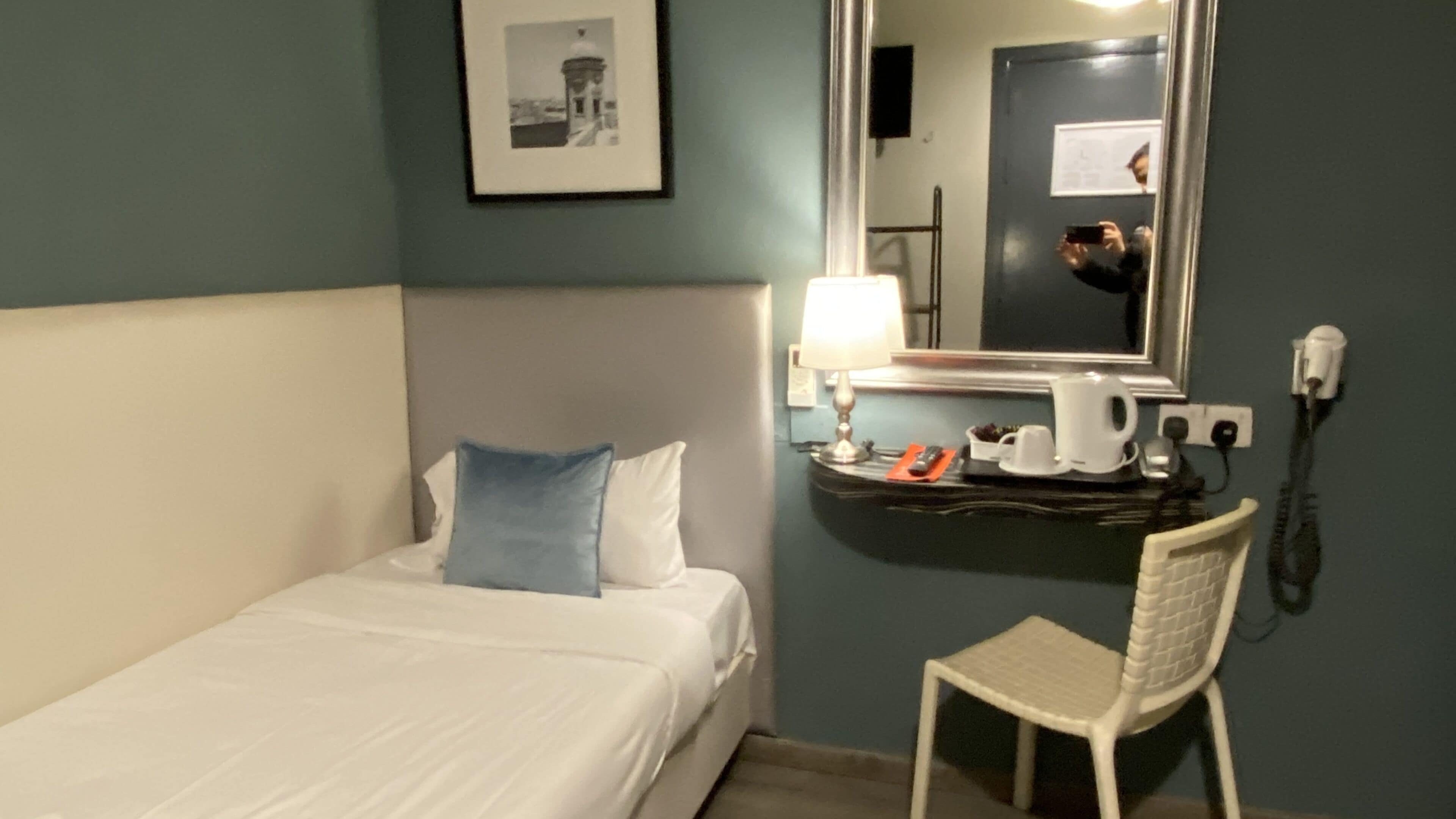 A hotel room with a bed, a chair, a desk with a lamp, a telephone, and a framed picture on the wall.