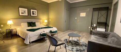 A hotel room with a bed, two bedside lamps, a chair, a small table, a sofa, and a door.