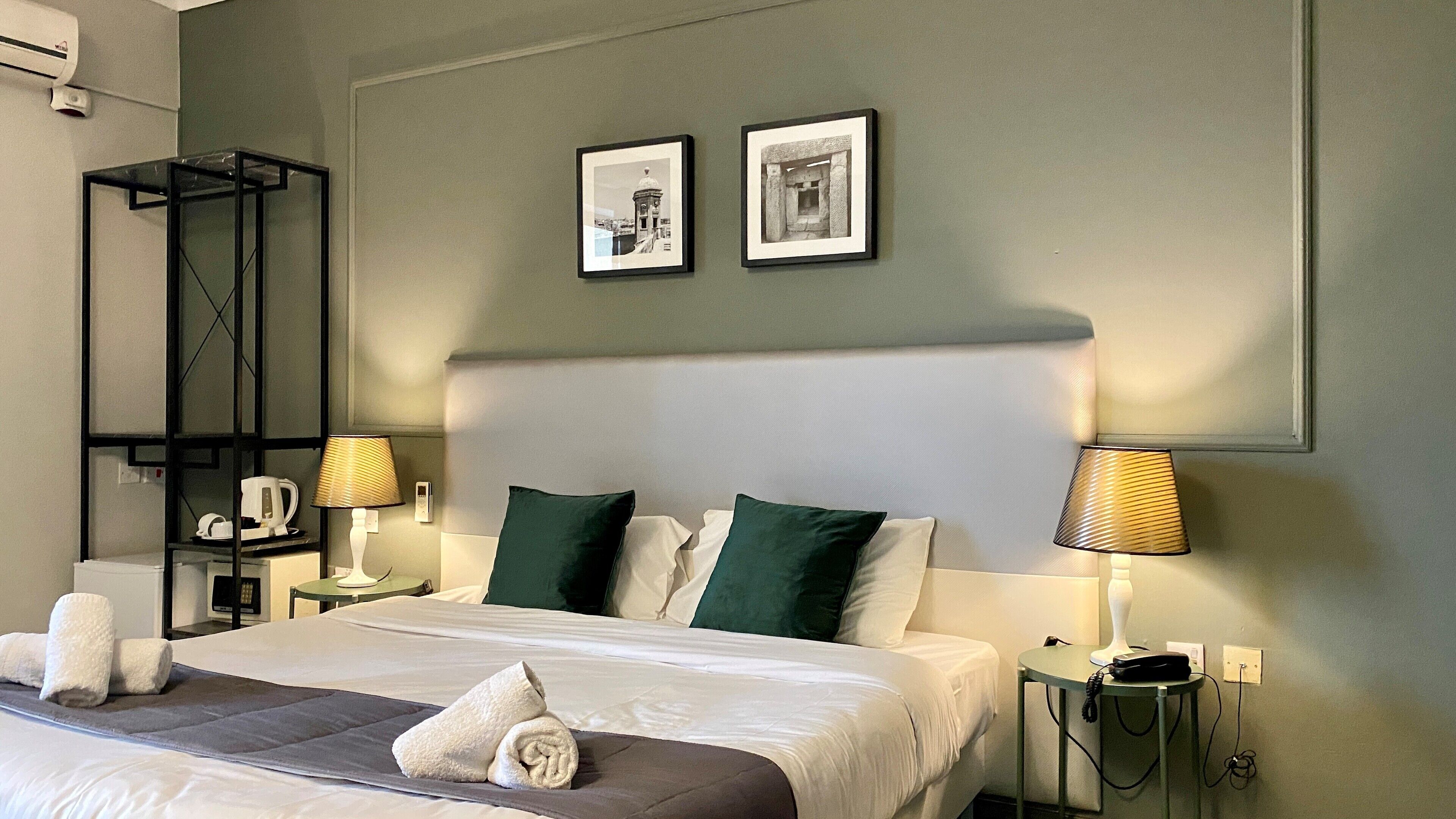 A hotel room with a large bed, two pillows, two bedside tables with lamps, a wall-mounted shelf, and two framed pictures.
