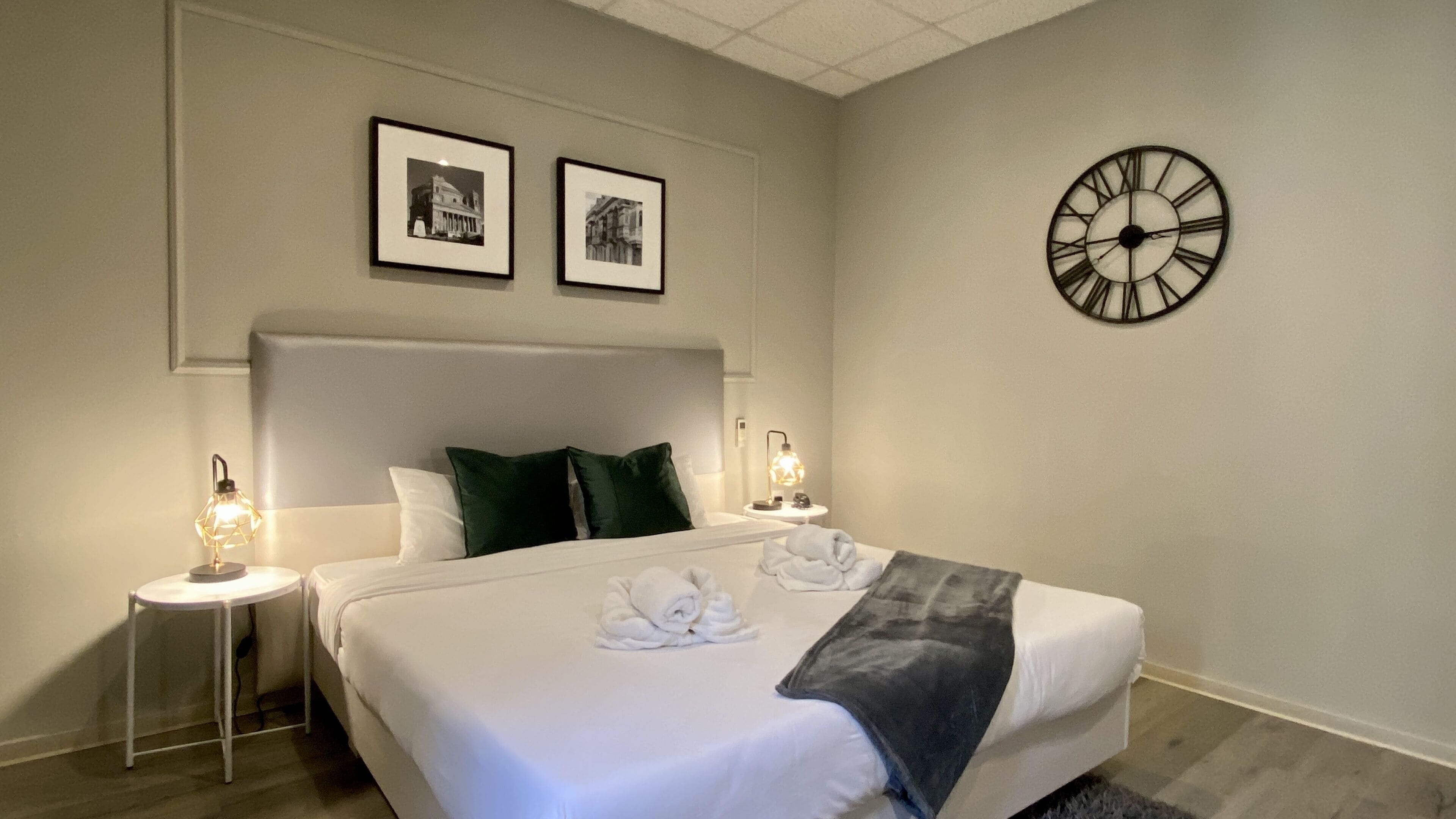 Un lit bien fait, avec du linge de lit blanc et deux oreillers, une horloge murale et deux tableaux encadrés au-dessus du lit.