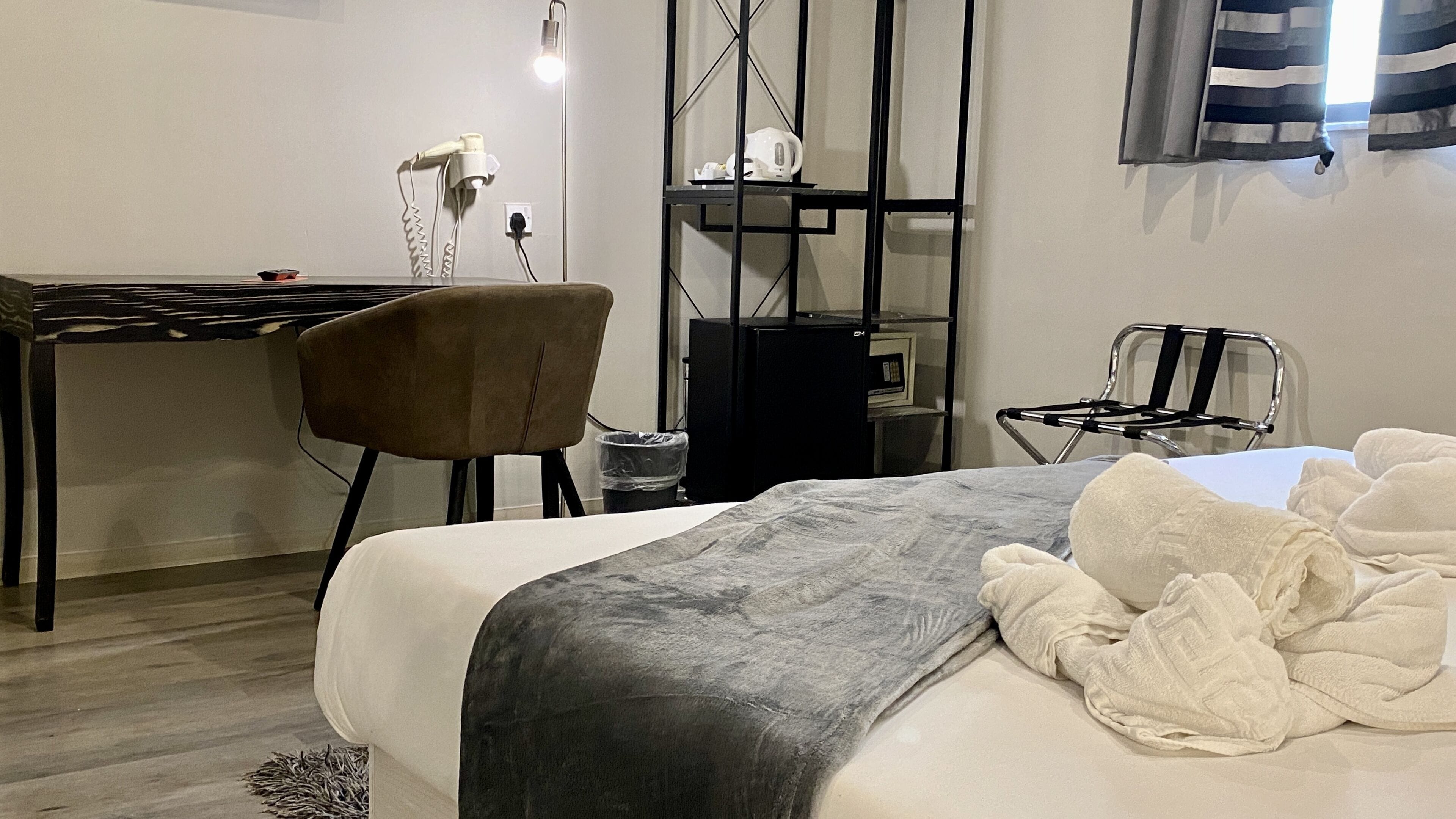 A hotel room with a bed, a desk, a chair, a small table, a shelf, and a window with curtains.