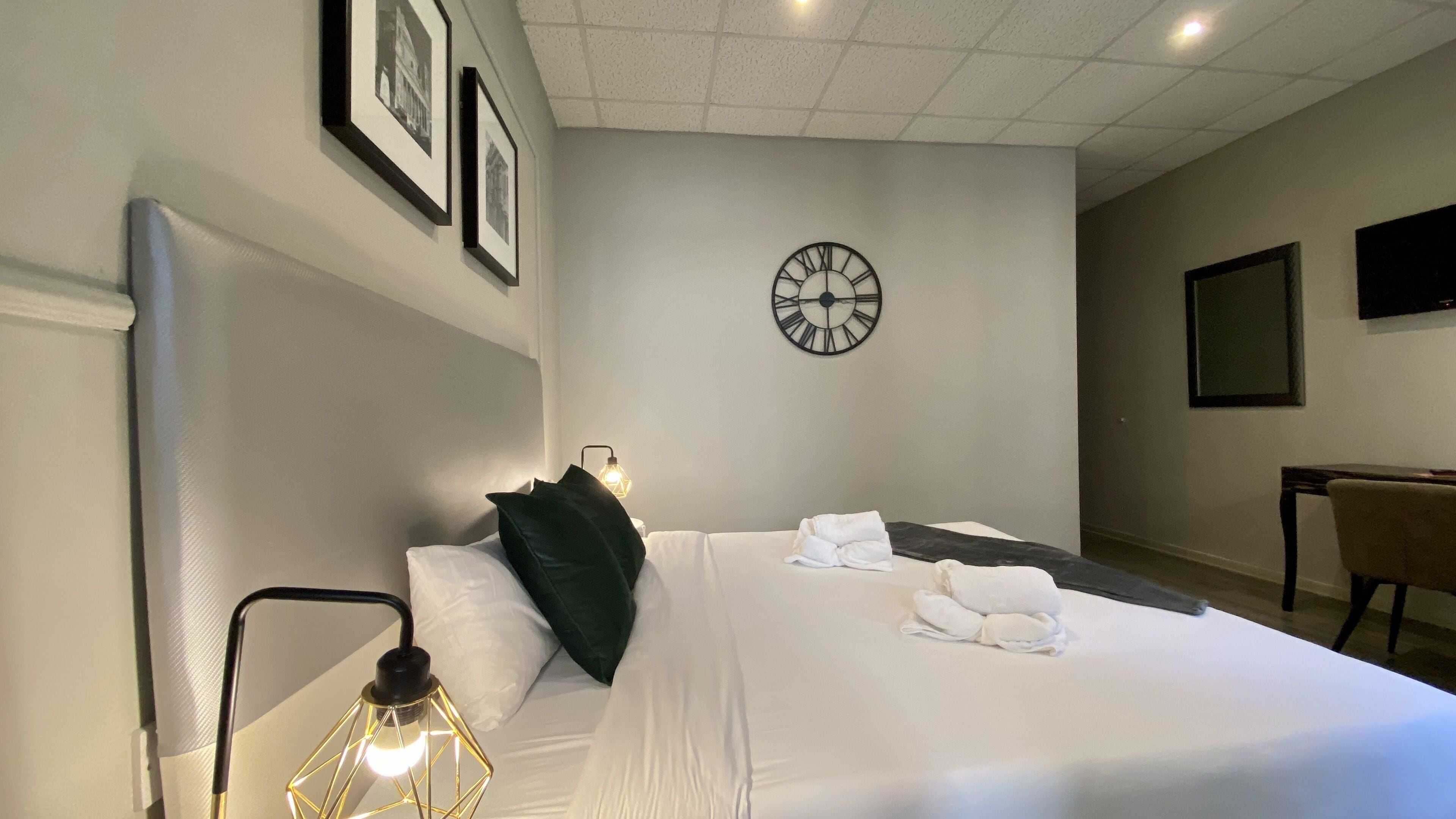 A neatly made bed with pillows, a bedside lamp, a clock on the wall, and a chair in the corner.