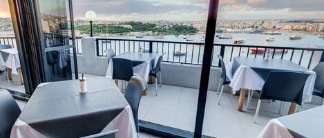 A balcony with tables and chairs, overlooking a harbor with boats and a cityscape.